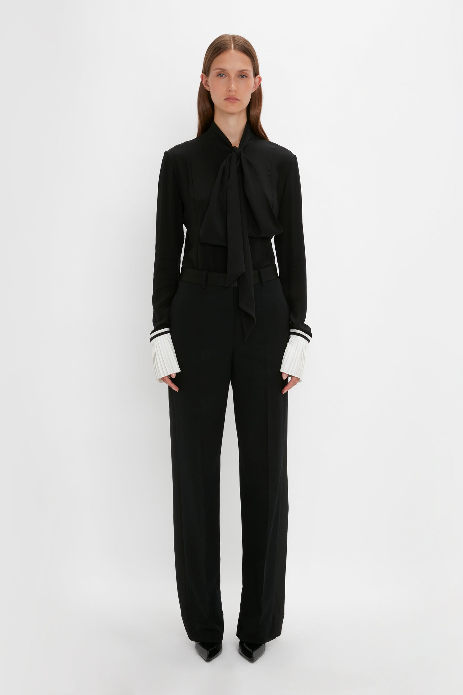 A person stands against a plain white background wearing the Pleat Cuff Blouse In Black by Victoria Beckham, staring directly ahead.