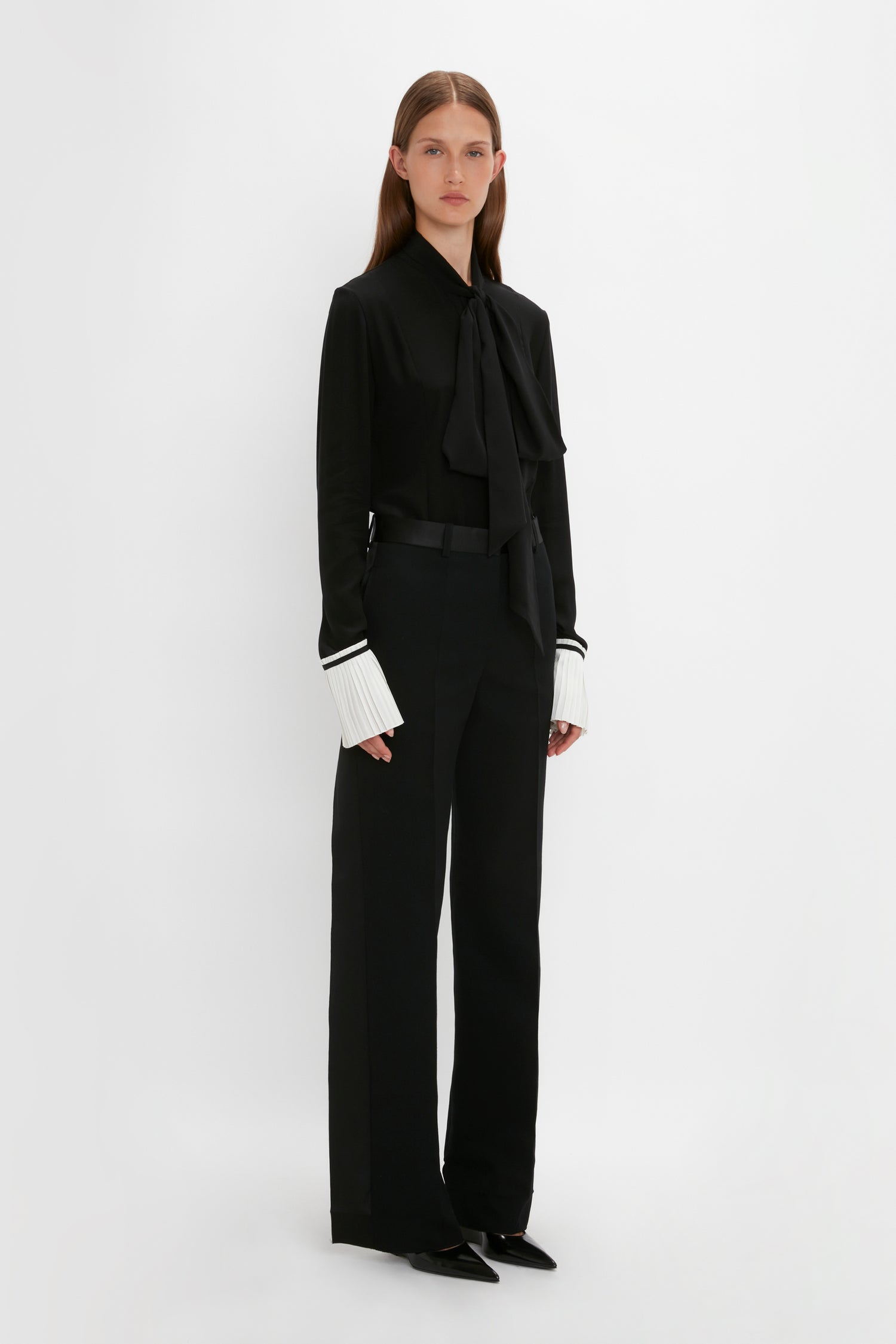 A person stands against a plain white background, wearing the Pleat Cuff Blouse In Black by Victoria Beckham, black trousers, and black pointed shoes.