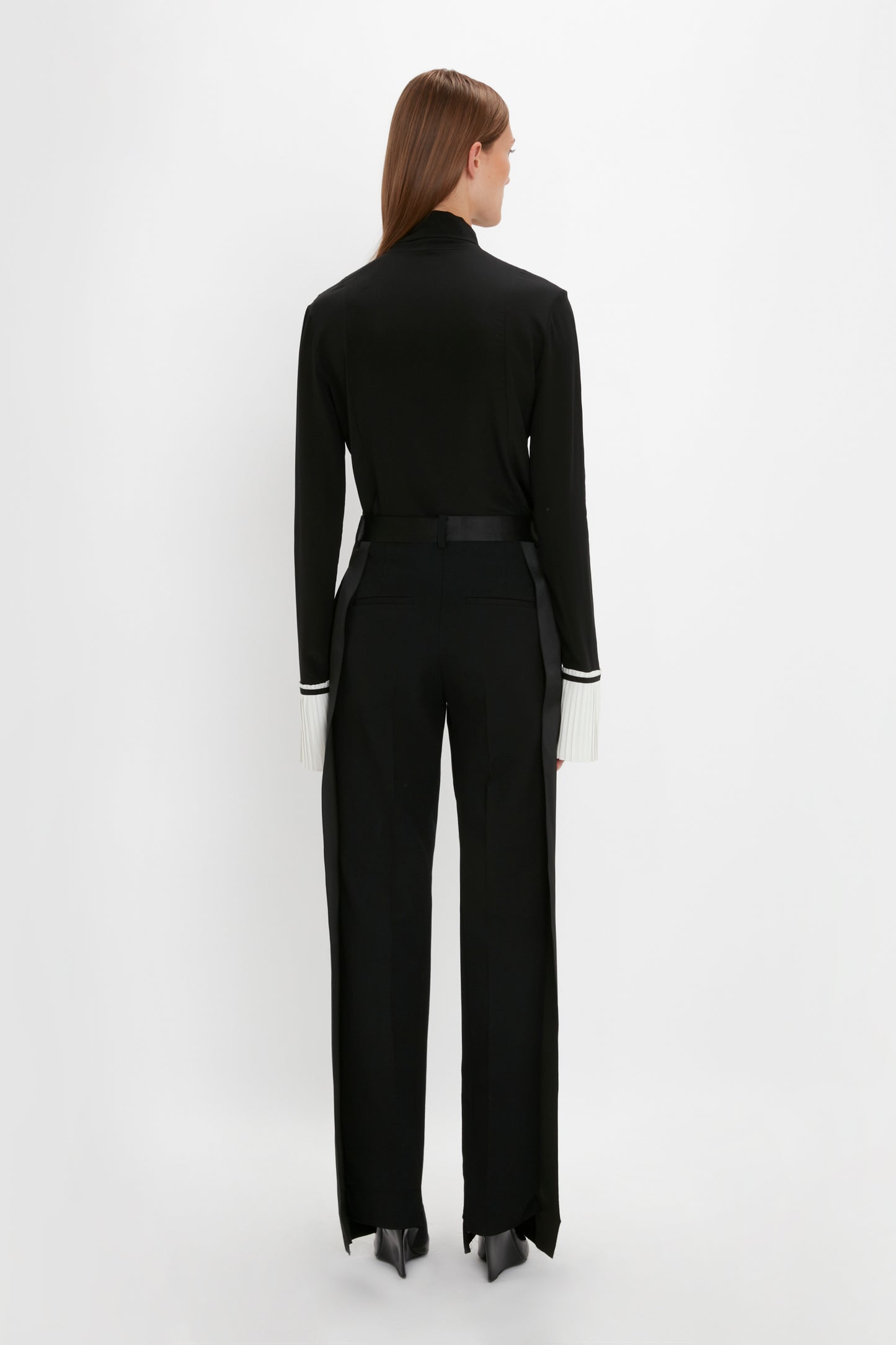 A person with long hair wearing a Pleat Cuff Blouse In Black by Victoria Beckham and black pants stands facing away against a white background, accented with knife pleat cuffs adding a touch of sophistication.
