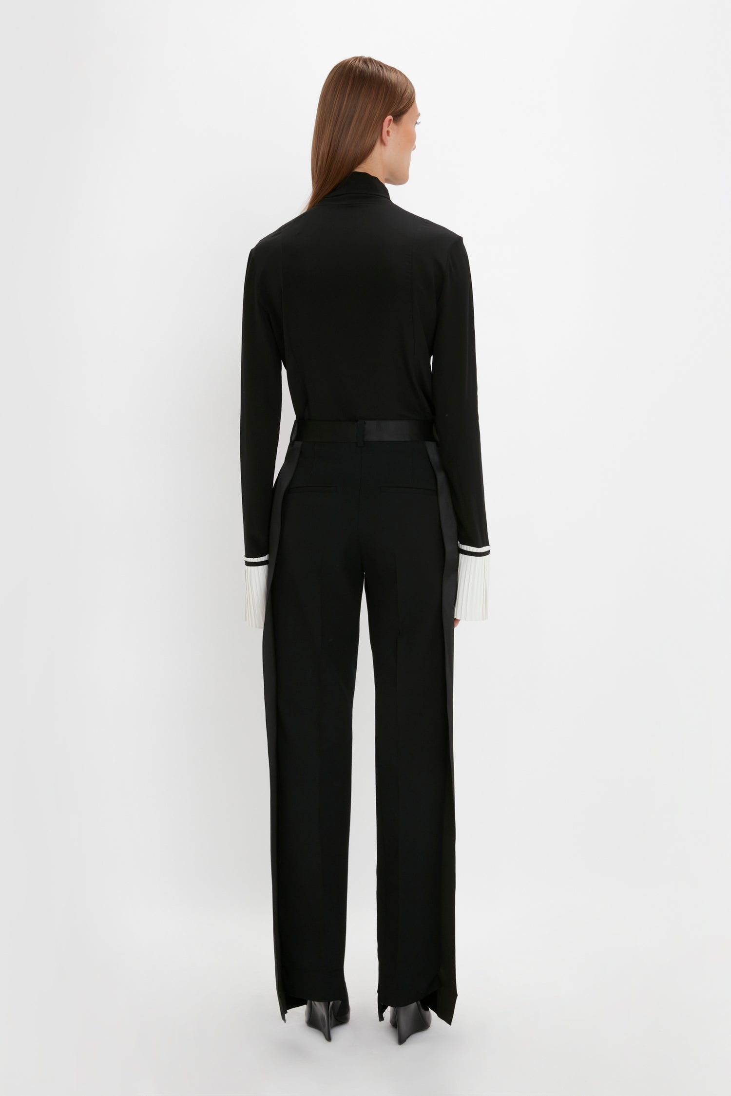 A person with long hair wearing a Pleat Cuff Blouse In Black by Victoria Beckham and black pants stands facing away against a white background, accented with knife pleat cuffs adding a touch of sophistication.