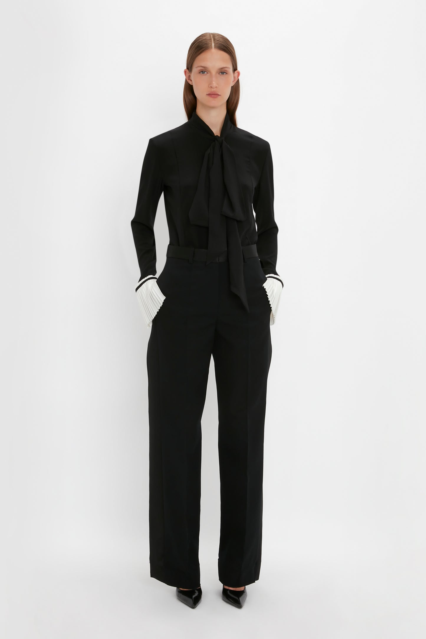 A woman stands upright against a plain white background, wearing a black outfit that features the Victoria Beckham Pleat Cuff Blouse In Black with a tie detail collar, wide-legged trousers, and pointed black shoes. Her long-sleeved top is elegant with knife pleat cuffs adding an extra touch of sophistication.