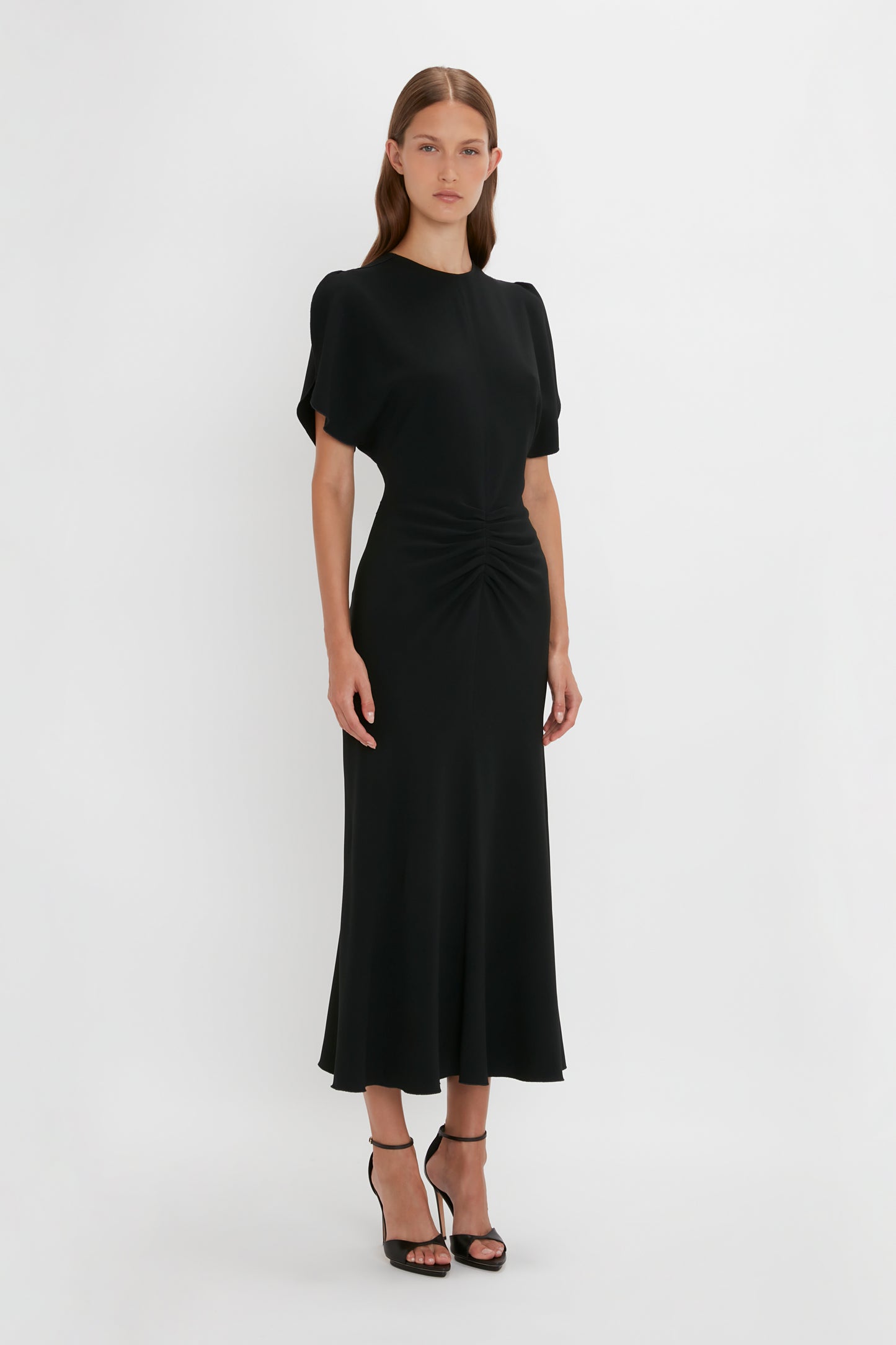 Woman in a Victoria Beckham black Gathered Waist Midi Dress with short sleeves and cinched waist detail, standing against a plain white background.