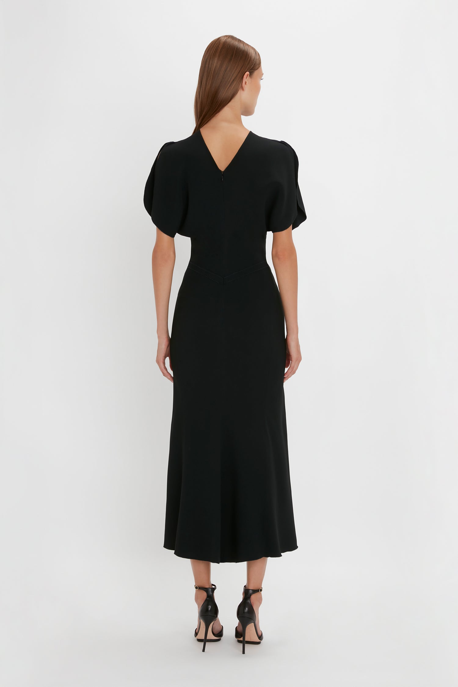A woman seen from behind, wearing a black, mid-calf length Gathered Waist Midi Dress in Black with short sleeves and standing against a white background.