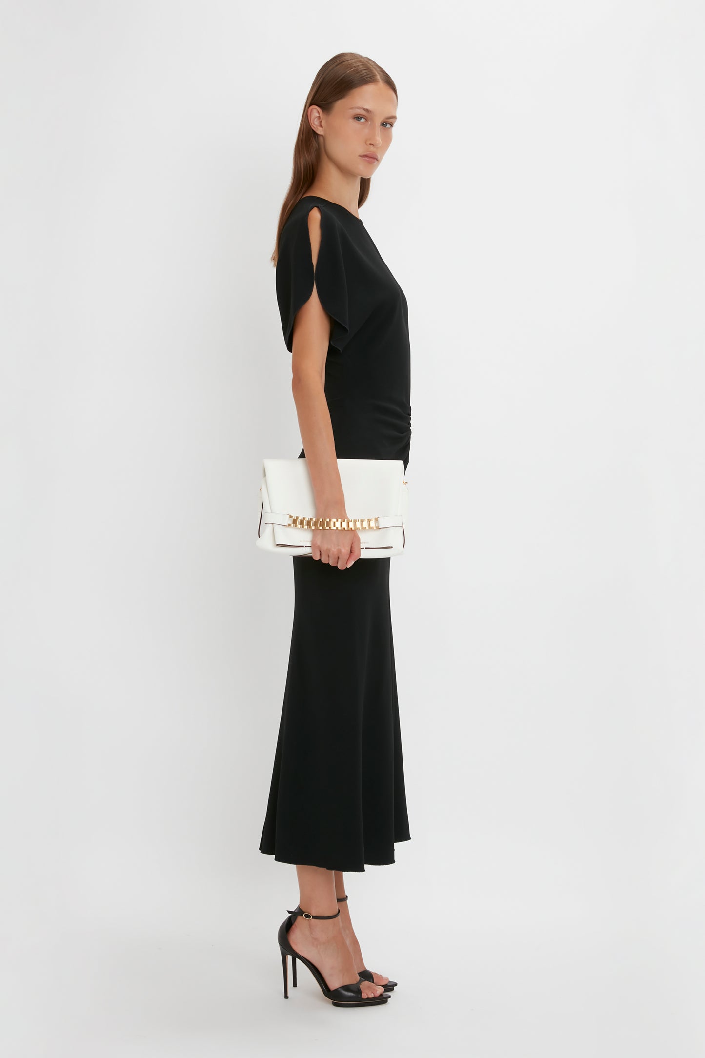 A woman in a black Victoria Beckham Gathered Waist Midi Dress and heels holds a white handbag, standing sideways against a white background.