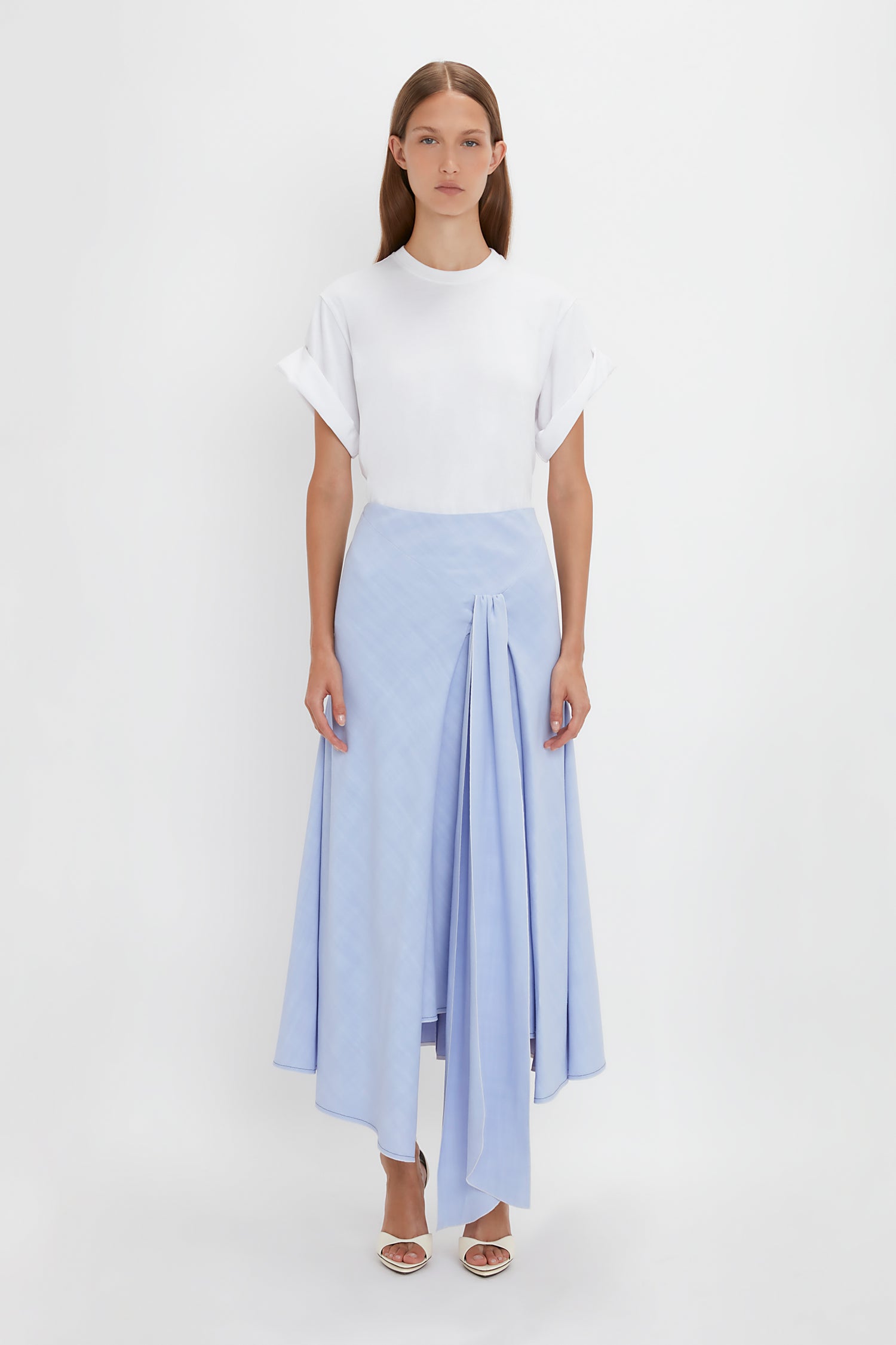 A person stands against a white background wearing a white short-sleeve top and an Asymmetric Tie Detail Skirt In Frost by Victoria Beckham in light blue fluid viscose crepe fabric, complemented by white open-toe sandals, showcasing modern femininity.