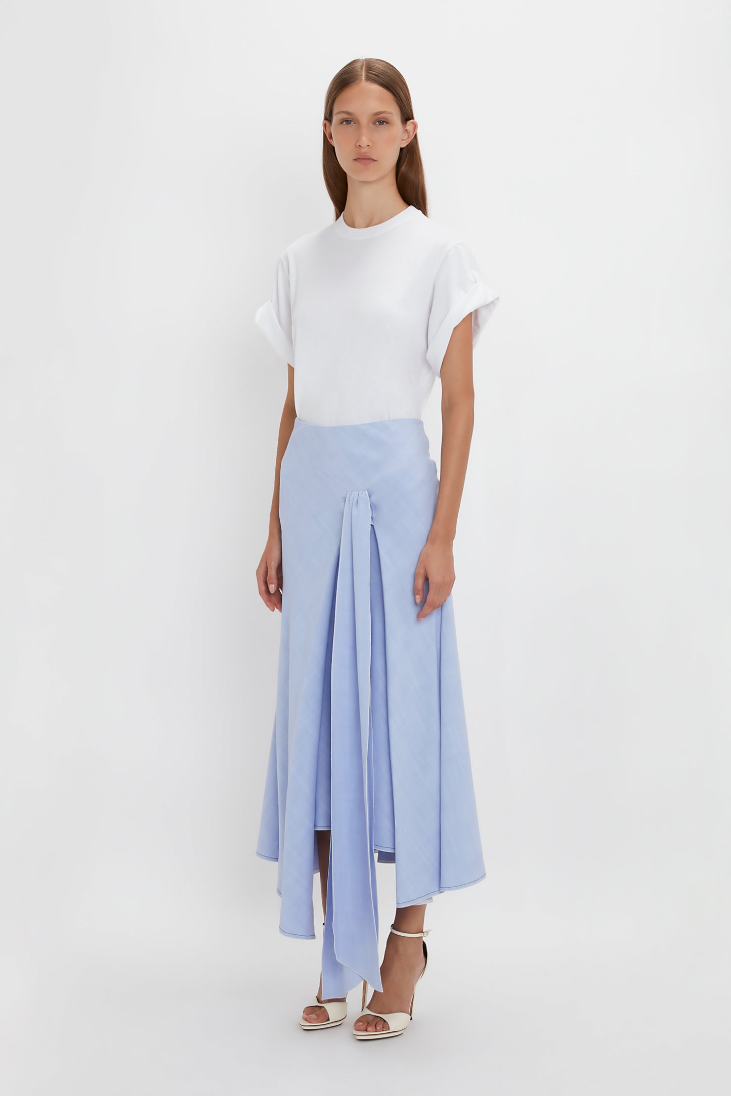 A woman stands against a plain white background, wearing a white t-shirt and Victoria Beckham's Asymmetric Tie Detail Skirt In Frost in fluid viscose crepe fabric. She is also wearing white open-toe heels, embodying modern femininity.