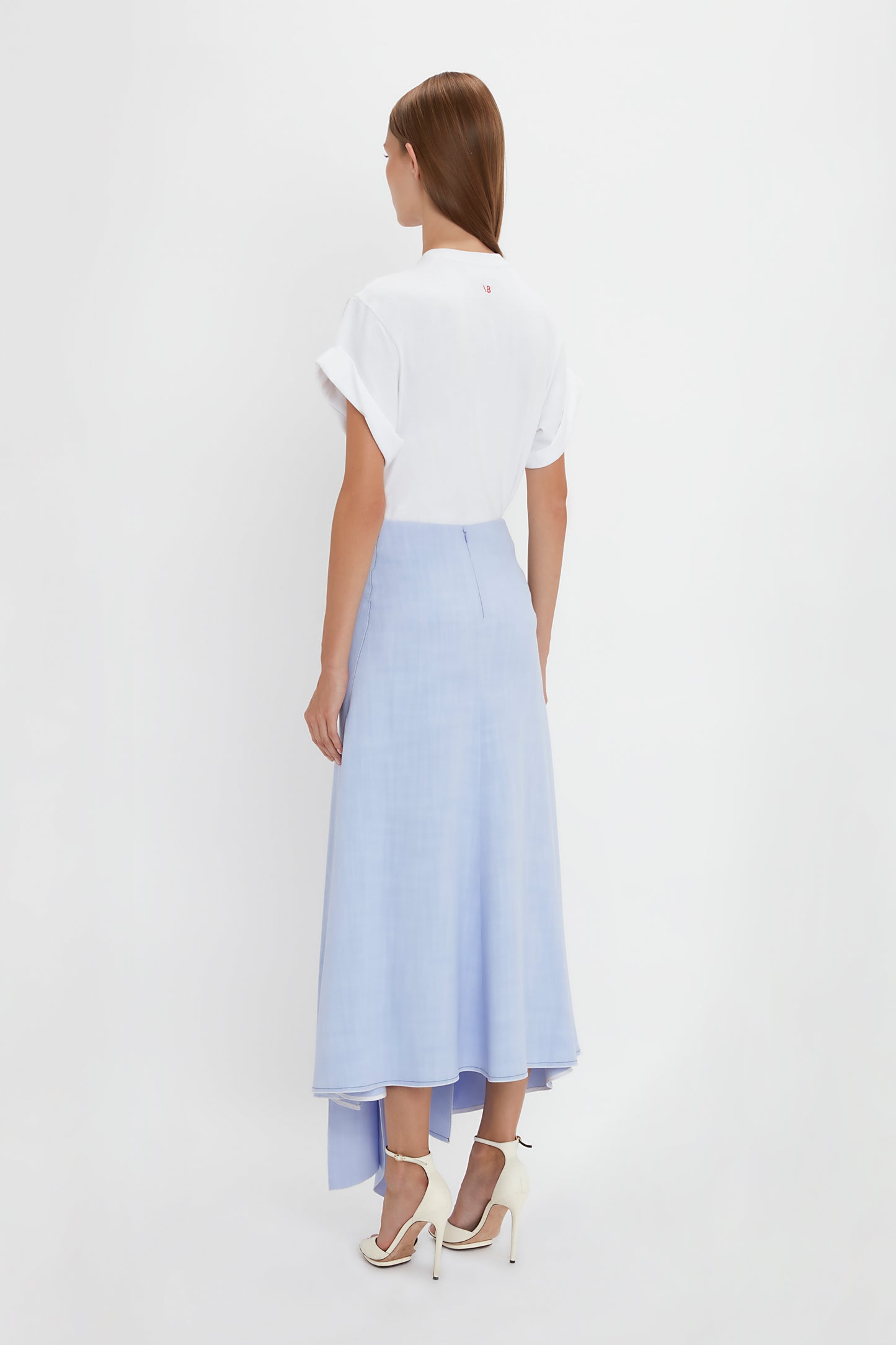 A woman is standing with her back to the camera, embodying modern femininity in a white short-sleeved top and a light blue Asymmetric Tie Detail Skirt In Frost by Victoria Beckham crafted from fluid viscose crepe fabric, paired with white high-heeled shoes against a pristine white background.