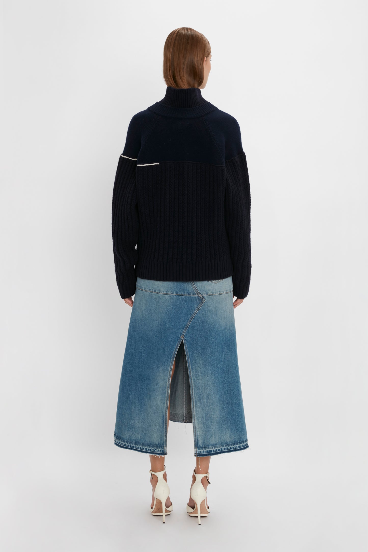 A person stands facing away, wearing a Victoria Beckham Collar Detail Jumper In Navy and a denim skirt with a front slit, paired with white high heels. The chunky knit adds texture and warmth to the look.