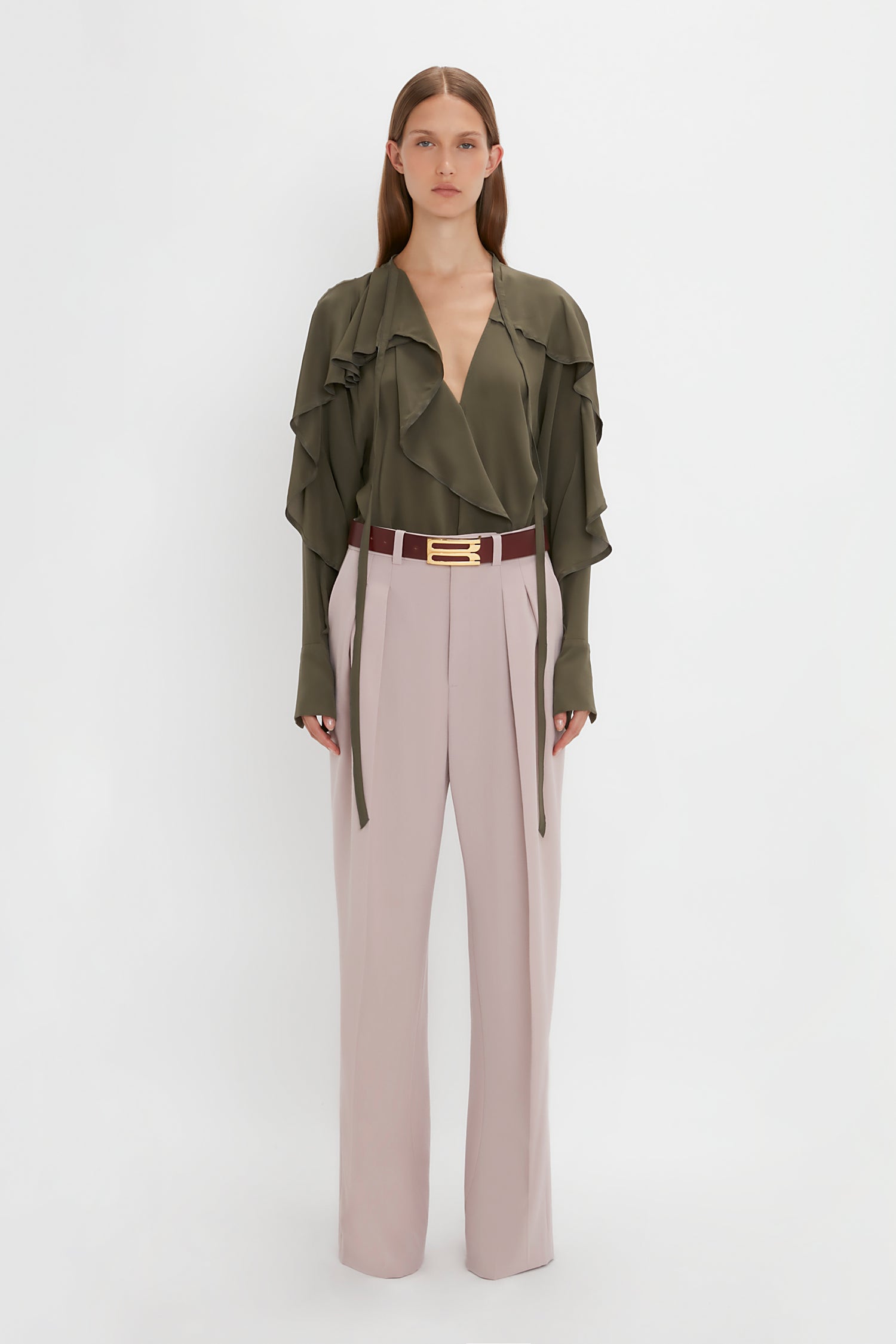 A person stands facing the camera wearing a green **Tie Detail Ruffle Blouse in Oregano by Victoria Beckham** and light pink Double Pleat Trousers with a brown belt. The background is plain white.