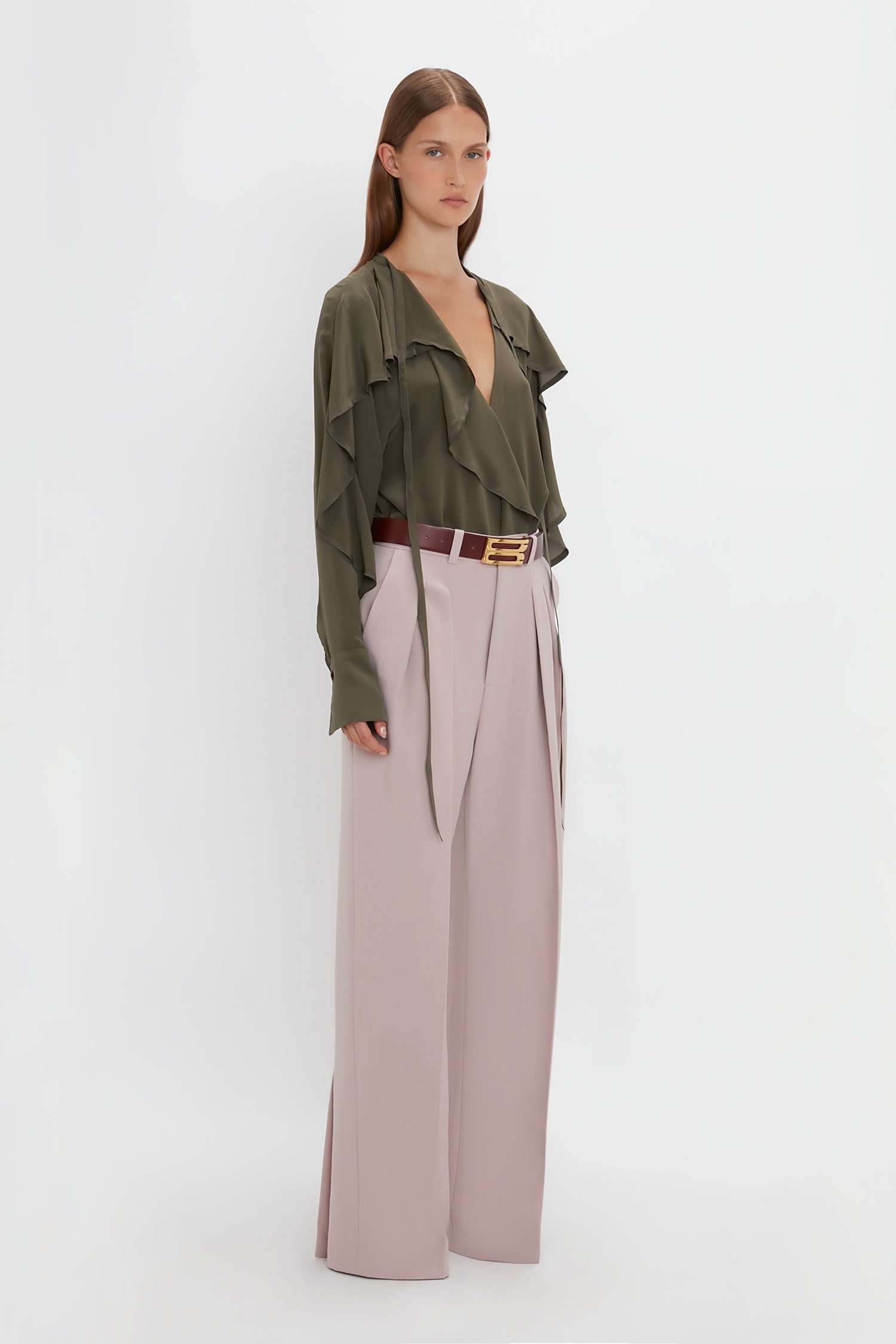 A person stands against a white background wearing a Victoria Beckham Tie Detail Ruffle Blouse in Oregano and high-waisted pale pink double pleat trousers, accessorized with a brown belt with a gold buckle.