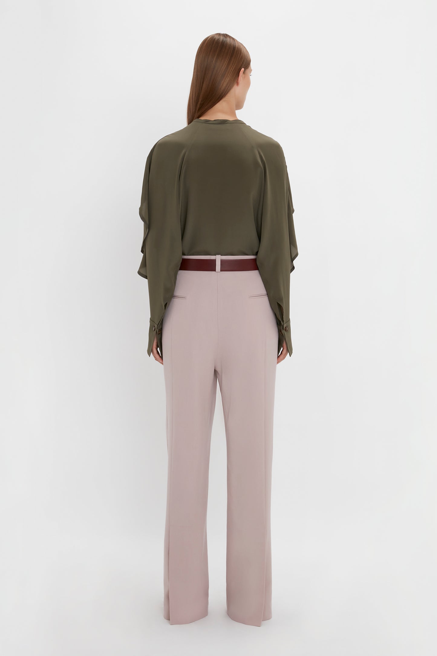A person with long hair is shown from the back, wearing a Victoria Beckham Tie Detail Ruffle Blouse in Oregano and light pink double pleat trousers with a red belt. They are standing against a plain white background.
