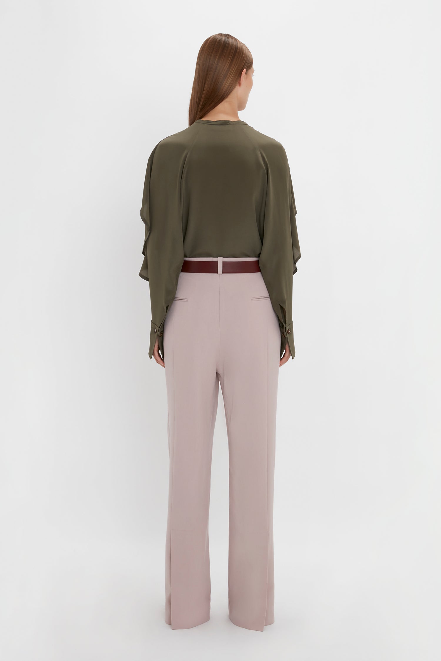 A person with long hair is shown from the back, wearing a Victoria Beckham Tie Detail Ruffle Blouse in Oregano and light pink double pleat trousers with a red belt. They are standing against a plain white background.