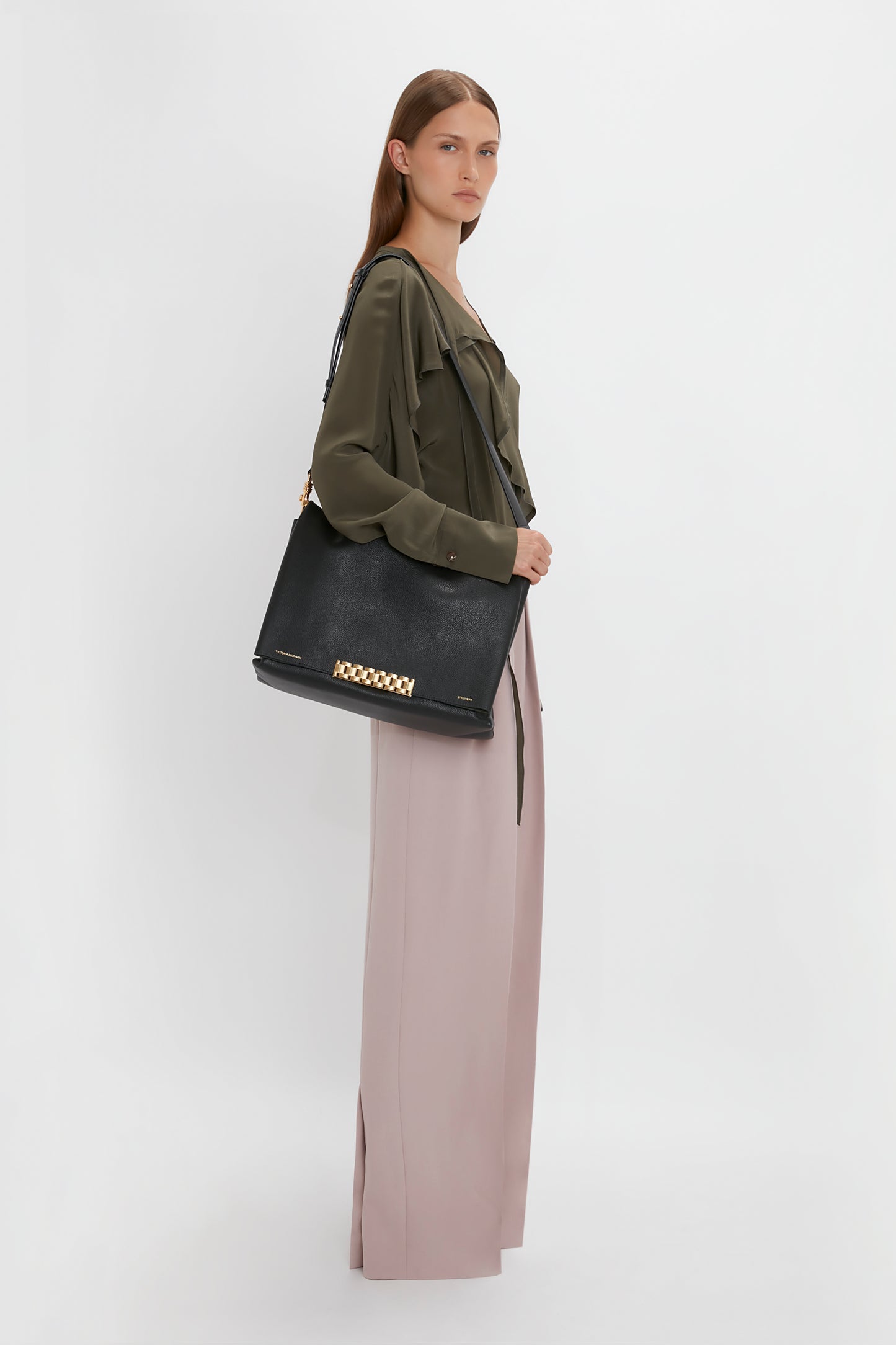 A person wearing a Tie Detail Ruffle Blouse in Oregano by Victoria Beckham and beige wide-leg pants stands in profile view, holding a black jumbo chain pouch with gold details.