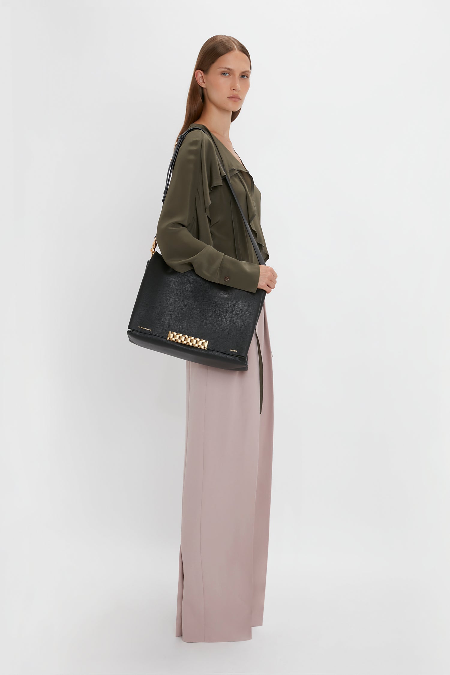 A person wearing a Tie Detail Ruffle Blouse in Oregano by Victoria Beckham and beige wide-leg pants stands in profile view, holding a black jumbo chain pouch with gold details.