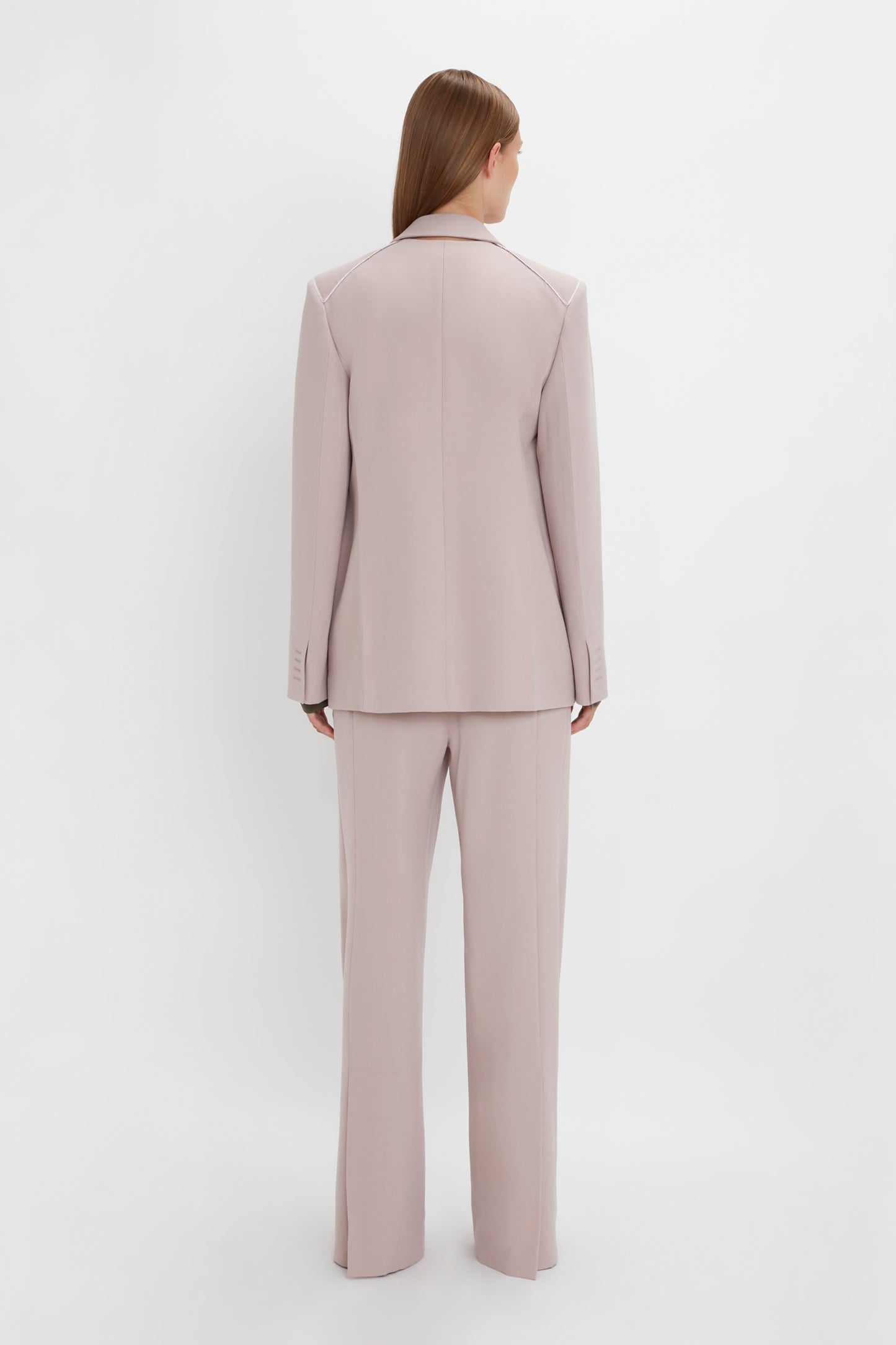 A person with long hair stands facing away, wearing a light pink, menswear-inspired pantsuit. The Double Panel Front Jacket In Rose Quartz by Victoria Beckham contrasts subtly against the plain white background.