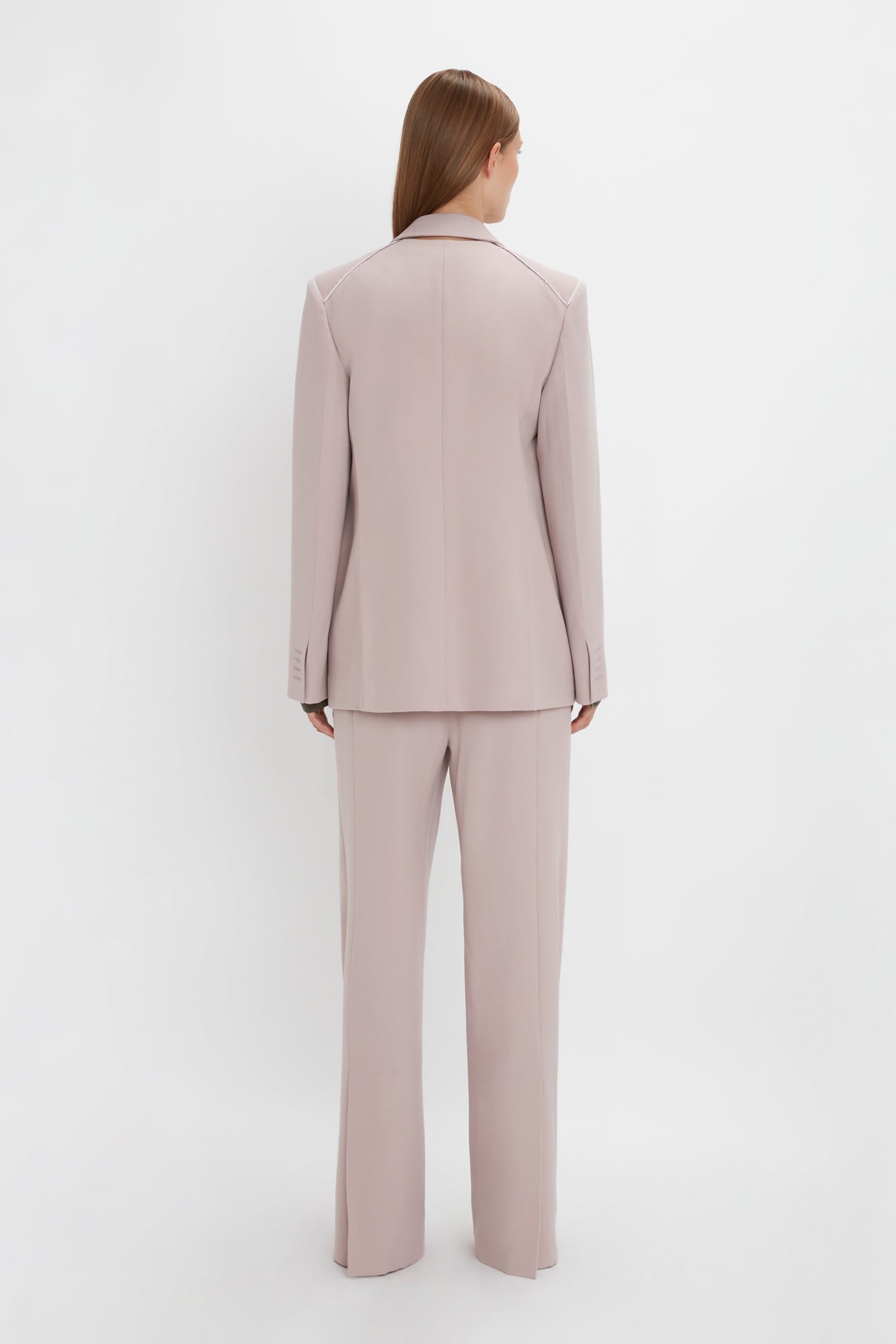 A person with long hair stands facing away, wearing a light pink, menswear-inspired pantsuit. The Double Panel Front Jacket In Rose Quartz by Victoria Beckham contrasts subtly against the plain white background.