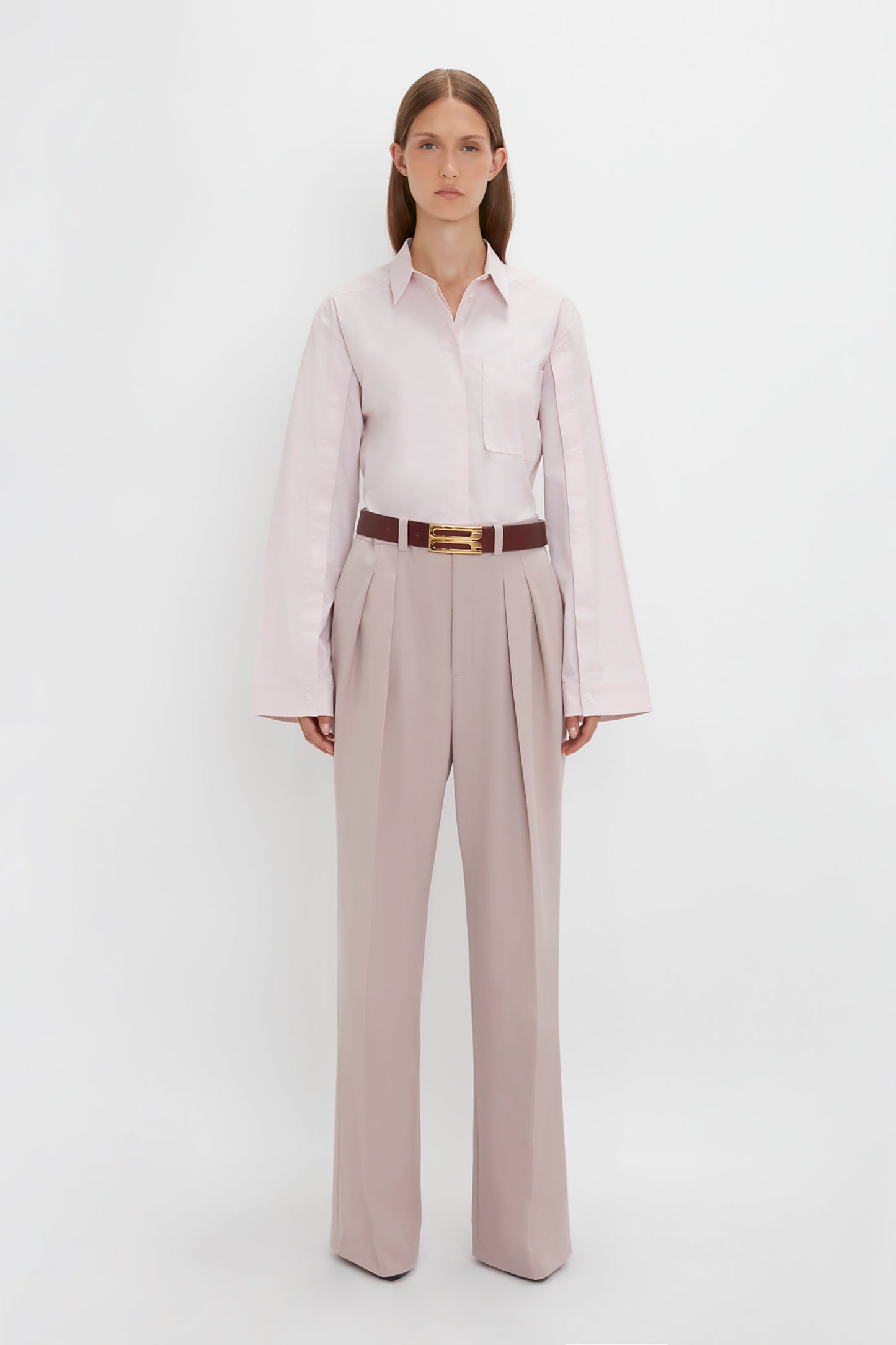 A person stands against a plain white background, wearing a light-colored Button Detail Cropped Shirt In Rose Quartz by Victoria Beckham and high-waisted, wide-leg trousers with a brown belt. Their arms are relaxed at their sides.