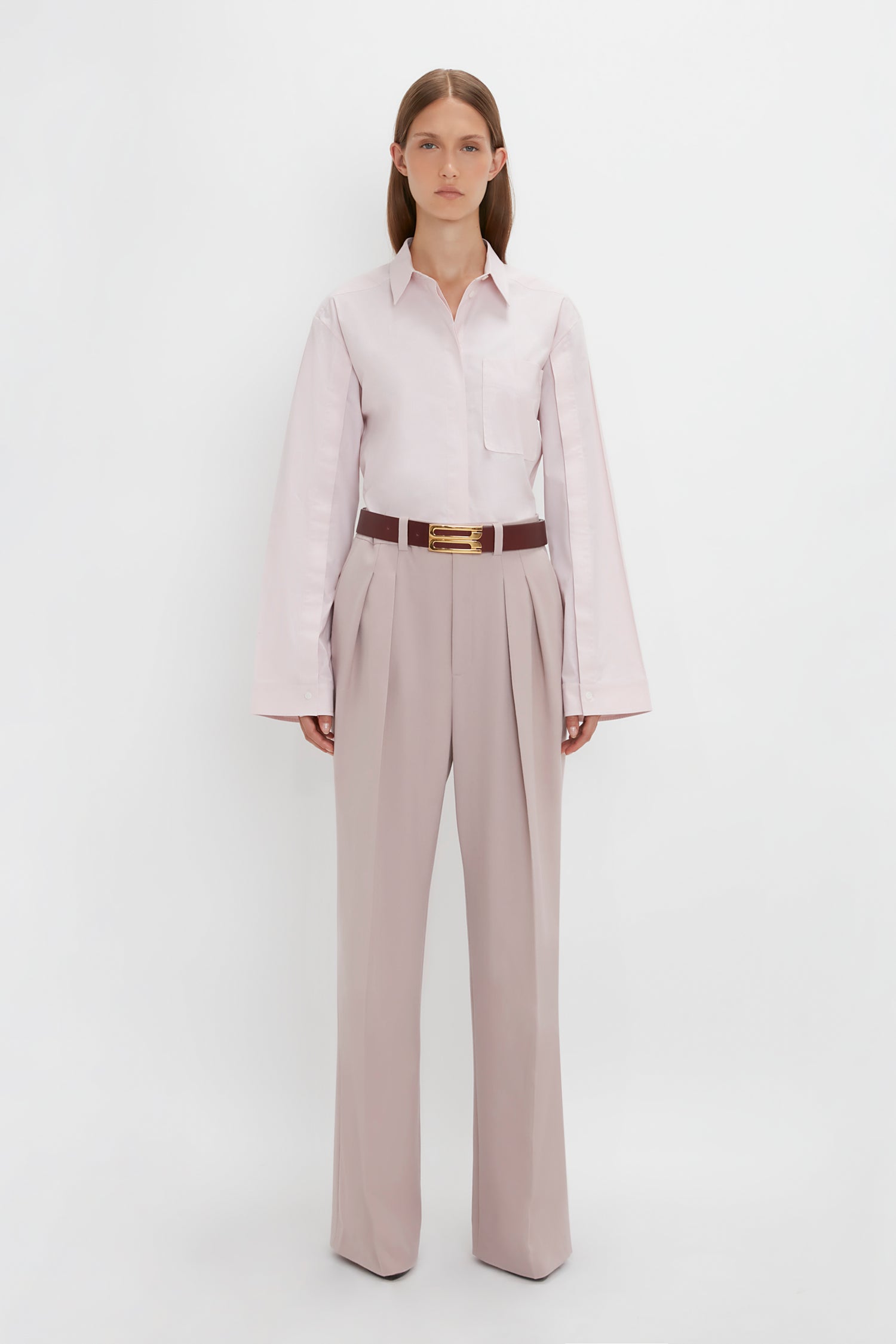 A person stands against a plain white background, wearing a light-colored Button Detail Cropped Shirt In Rose Quartz by Victoria Beckham and high-waisted, wide-leg trousers with a brown belt. Their arms are relaxed at their sides.