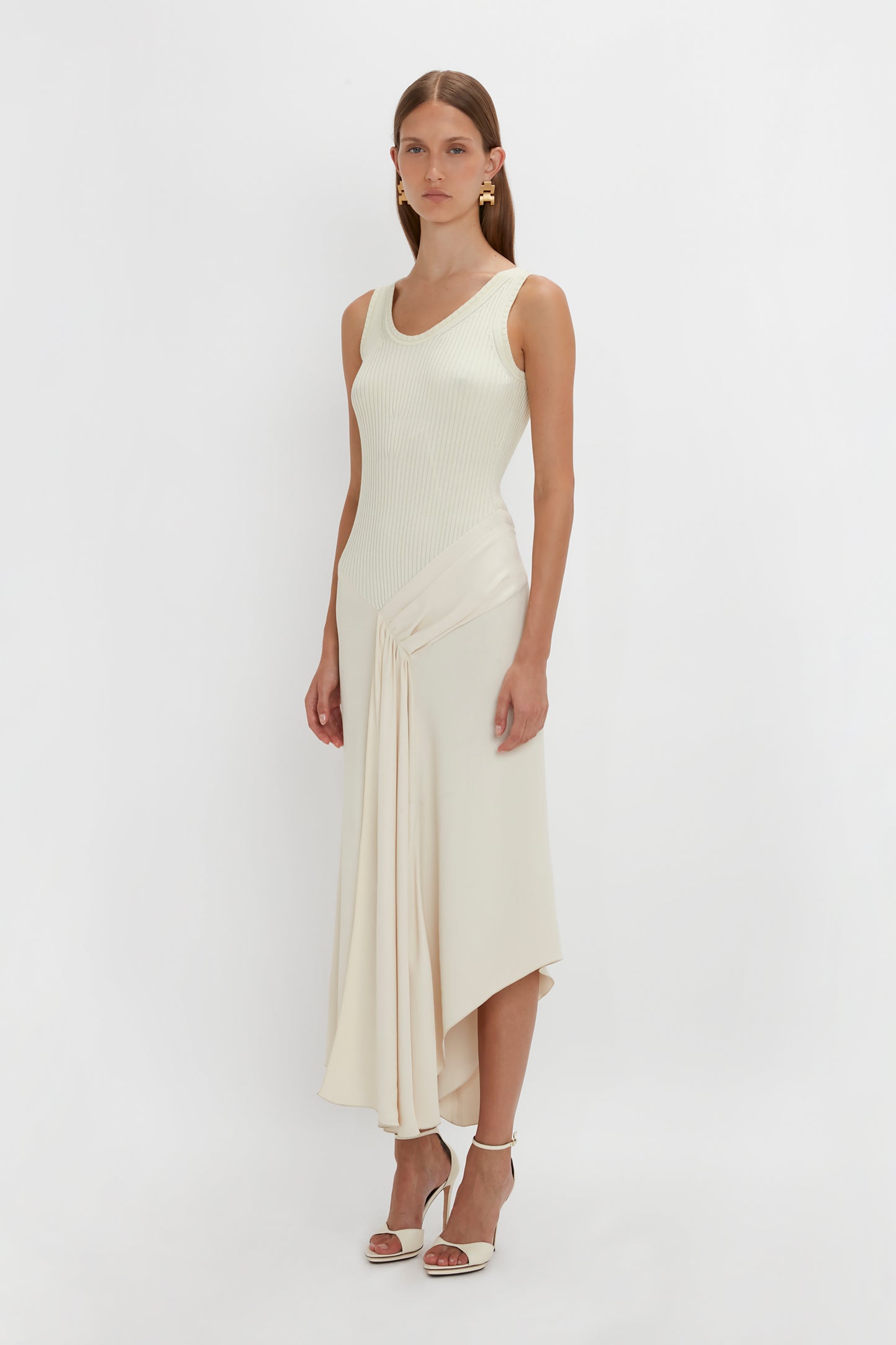 A woman in a Sleeveless Tie Detail Dress In Cream by Victoria Beckham stands against a plain white background, wearing white high-heeled sandals and gold earrings.