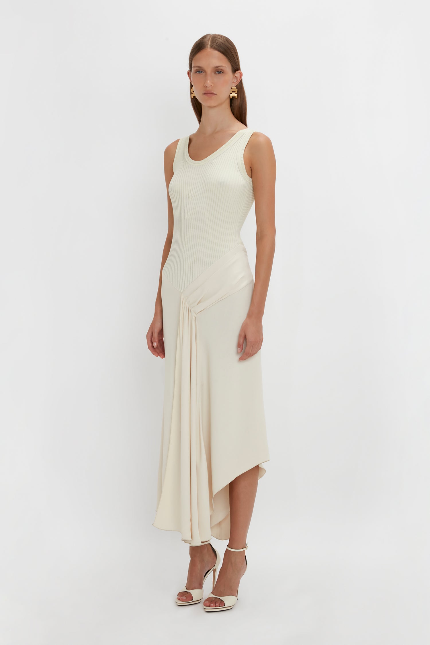 A woman in a Sleeveless Tie Detail Dress In Cream by Victoria Beckham stands against a plain white background, wearing white high-heeled sandals and gold earrings.