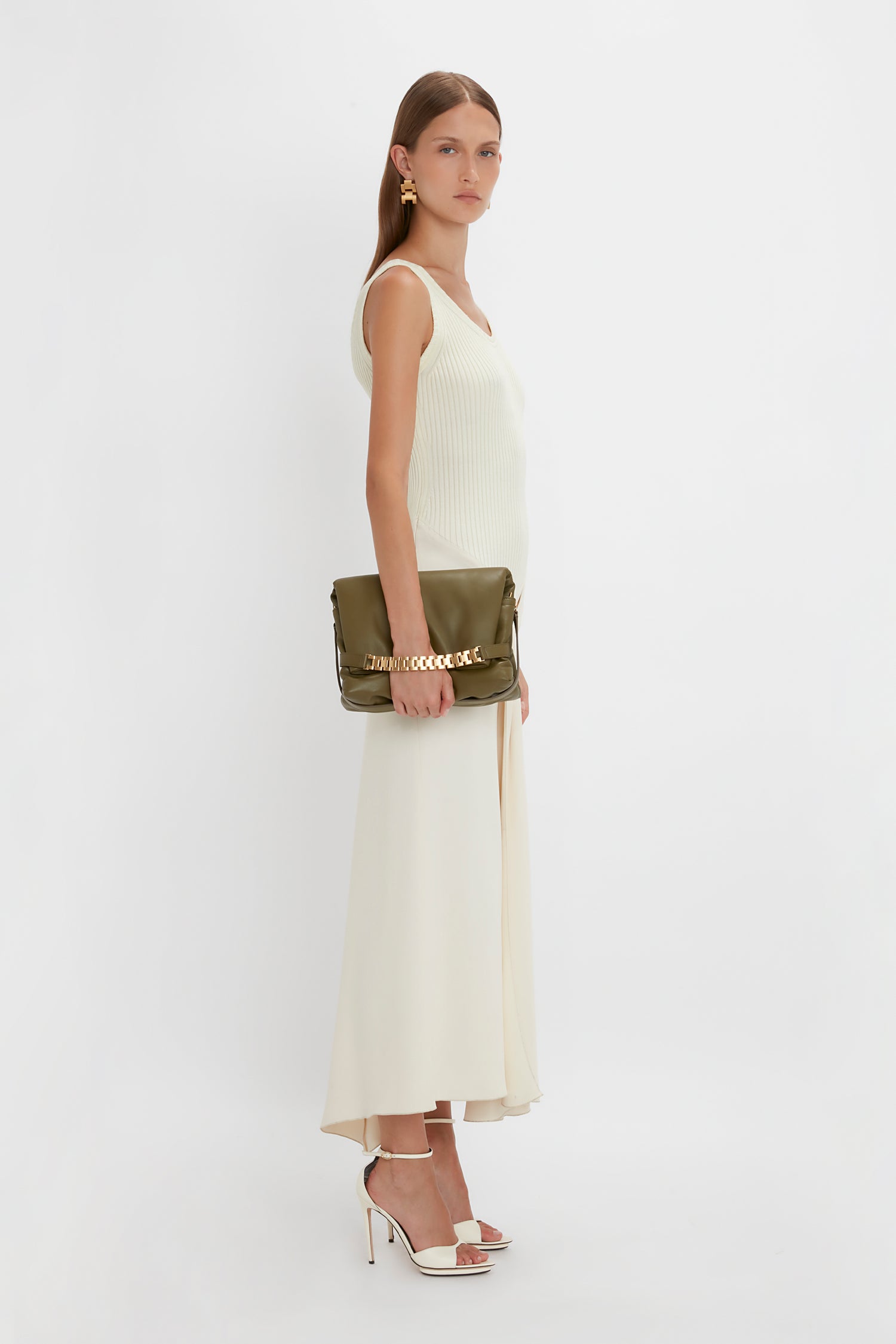 A woman stands against a plain backdrop, wearing a Victoria Beckham Sleeveless Tie Detail Dress In Cream with an asymmetric hemline, white high-heeled sandals, and holding a metallic green clutch. She has long brown hair and is wearing gold earrings.