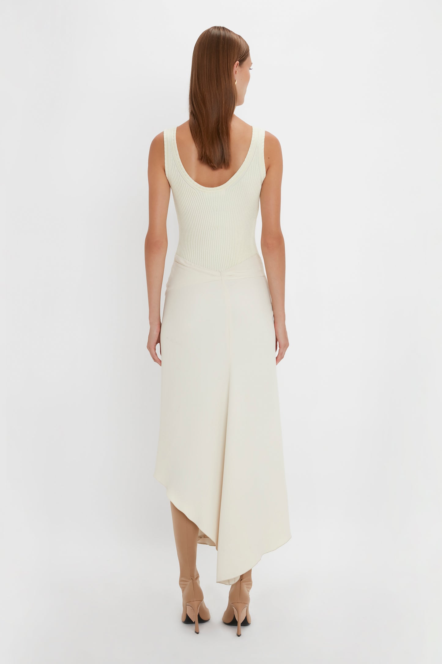 A woman with long brown hair is standing with her back to the camera. She is wearing a Sleeveless Tie Detail Dress In Cream by Victoria Beckham, paired with beige high heels. The background is plain white.