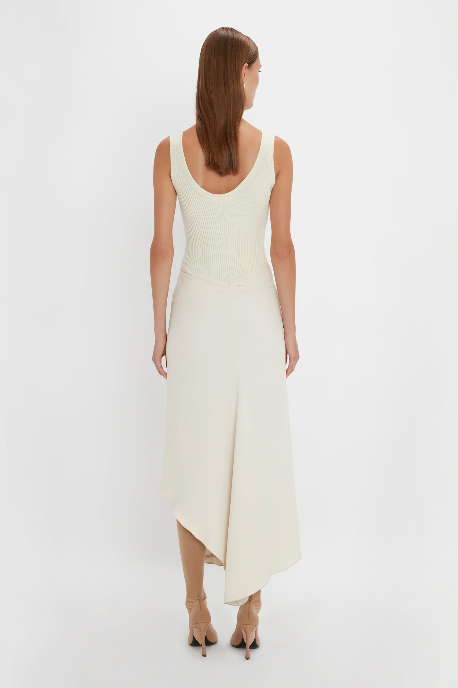 A woman with long brown hair is standing with her back to the camera. She is wearing a Sleeveless Tie Detail Dress In Cream by Victoria Beckham, paired with beige high heels. The background is plain white.