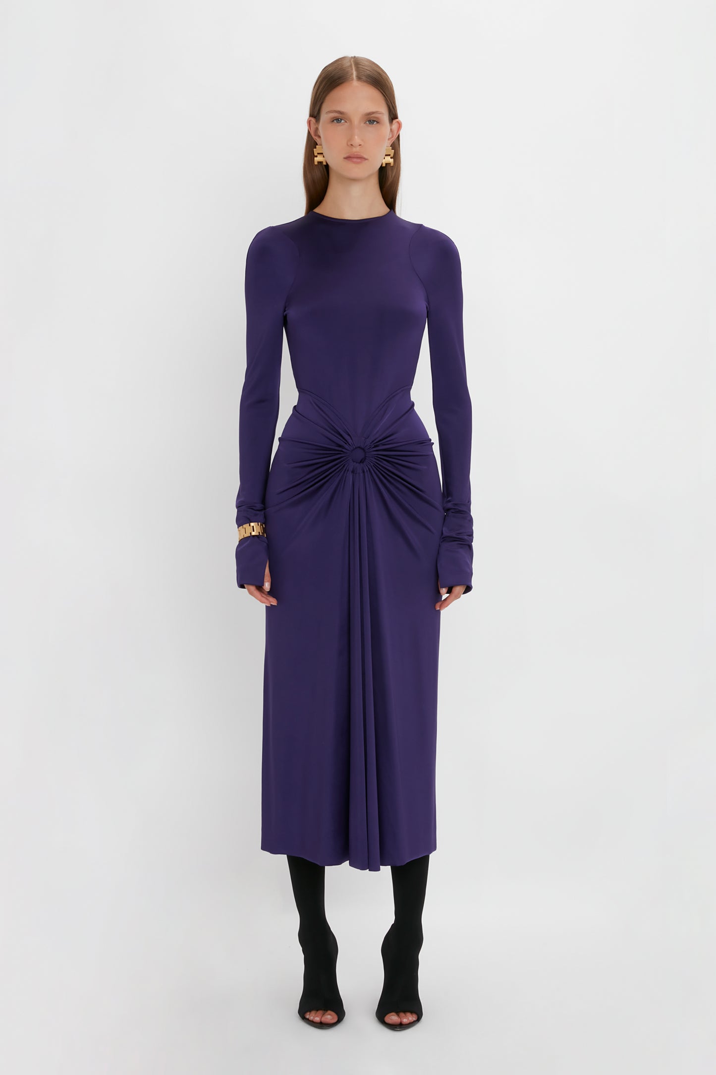 A person stands against a white background wearing a sensuous and playful Victoria Beckham Long Sleeve Gathered Midi Dress In Ultraviolet, made from body-sculpting stretch fabric. The ensemble is completed with black tights and open-toe black shoes, perfectly highlighting the gathered center detail.