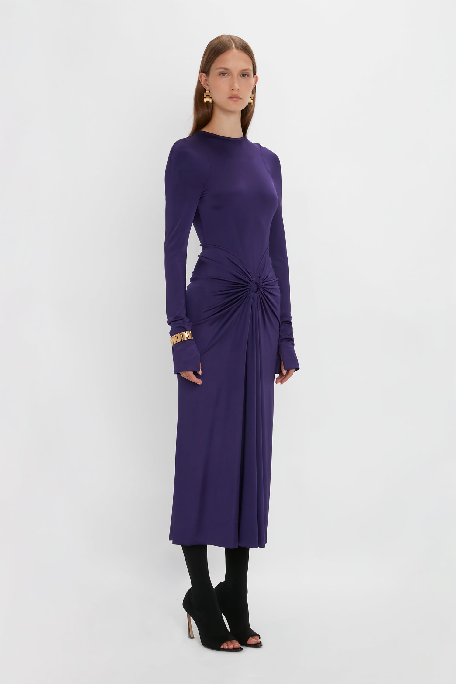 A woman with long hair stands against a white background wearing a sensuous and playful, Long Sleeve Gathered Midi Dress In Ultraviolet by Victoria Beckham. She is also wearing black open-toe heels.