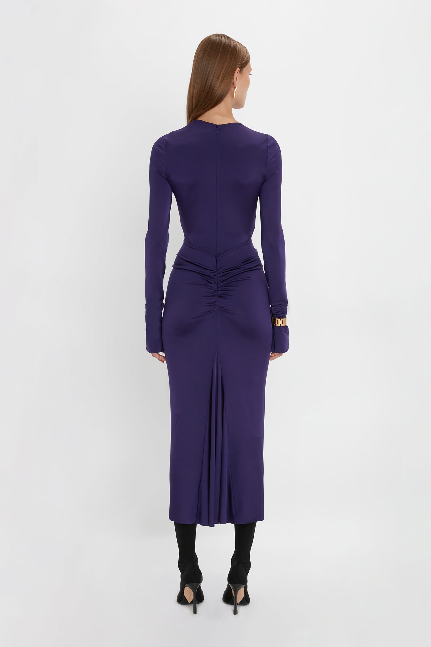 A person stands facing away, wearing a Victoria Beckham Long Sleeve Gathered Midi Dress In Ultraviolet with ruched detailing at the back and a rear slit, showcasing the sensuous and playful design. Paired with black heeled shoes, it's made from body-sculpting stretch fabric for a flawless fit.
