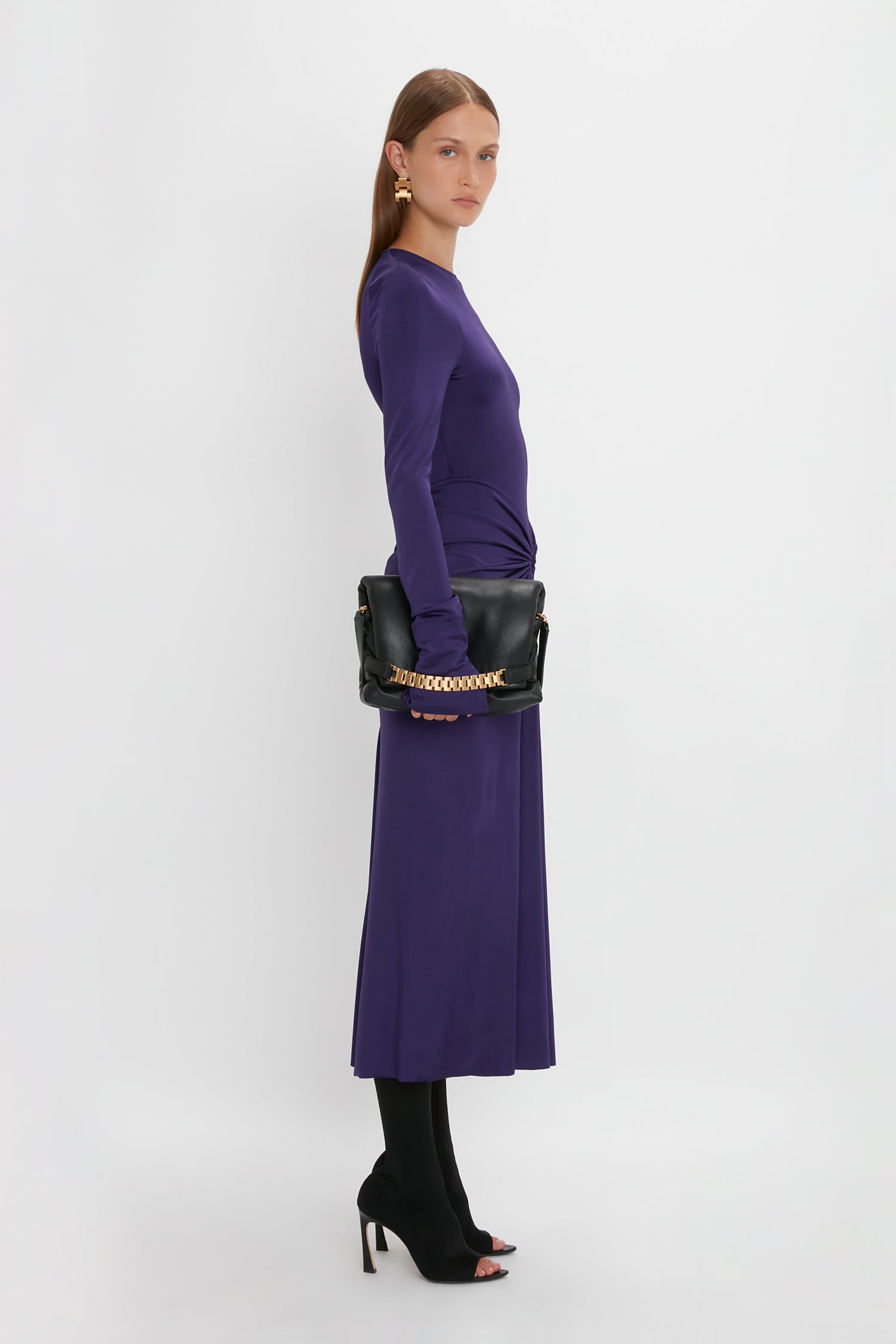 A sensuous and playful woman with long hair, wearing the Victoria Beckham Long Sleeve Gathered Midi Dress In Ultraviolet made of body-sculpting stretch fabric and black heels, holds a black handbag with a gold chain, posing against a plain white background.