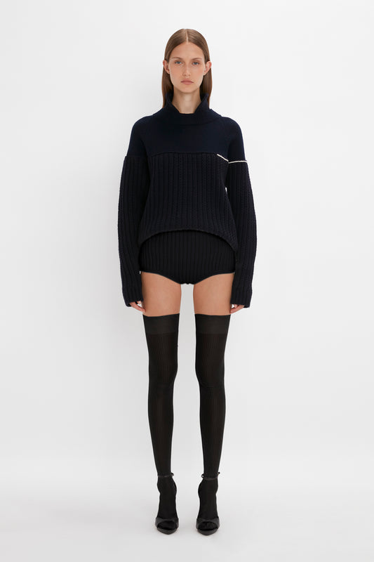 A woman stands center in a studio, wearing a black ribbed sweater dress, Victoria Beckham Exclusive Over The Knee Socks In Black, and short boots, facing forward with a neutral expression.