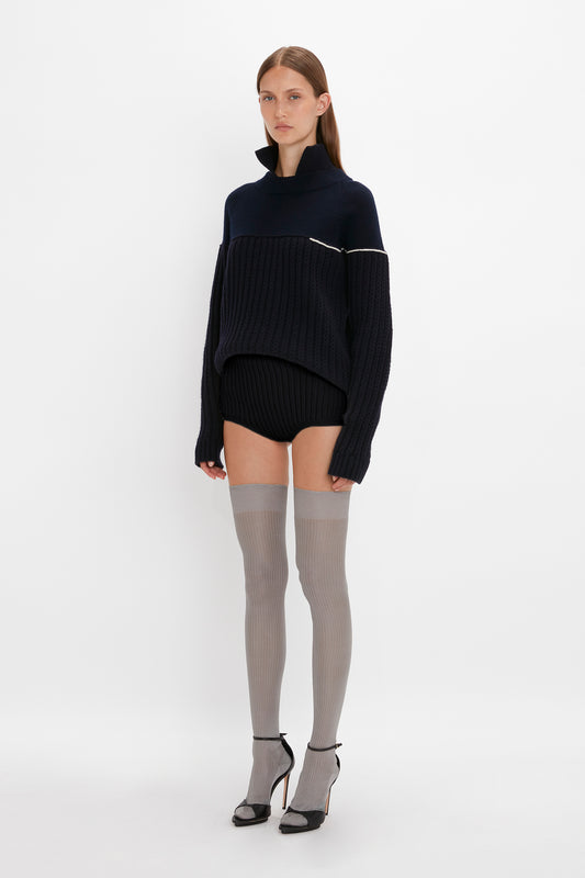 A model stands against a white background wearing a navy blue oversized sweater, silk Exclusive Over The Knee Socks In Grey, and black high heels by Victoria Beckham.