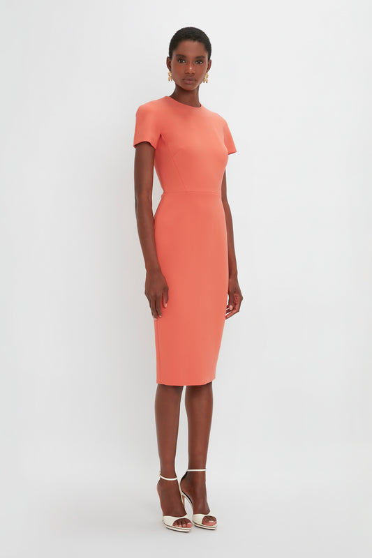 A woman in a fitted t-shirt dress in papaya from Victoria Beckham and gold sandals stands against a white background.