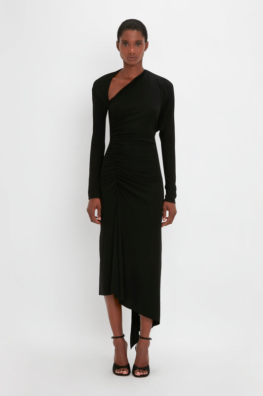 A woman in a Victoria Beckham chic black asymmetrical dress with an asymmetric cut-out and black strappy heels stands against a plain white background.