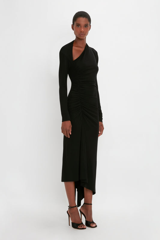 A black woman in a Victoria Beckham Slash-Neck Ruched Midi Dress In Black with an asymmetric cut-out poses against a white background.