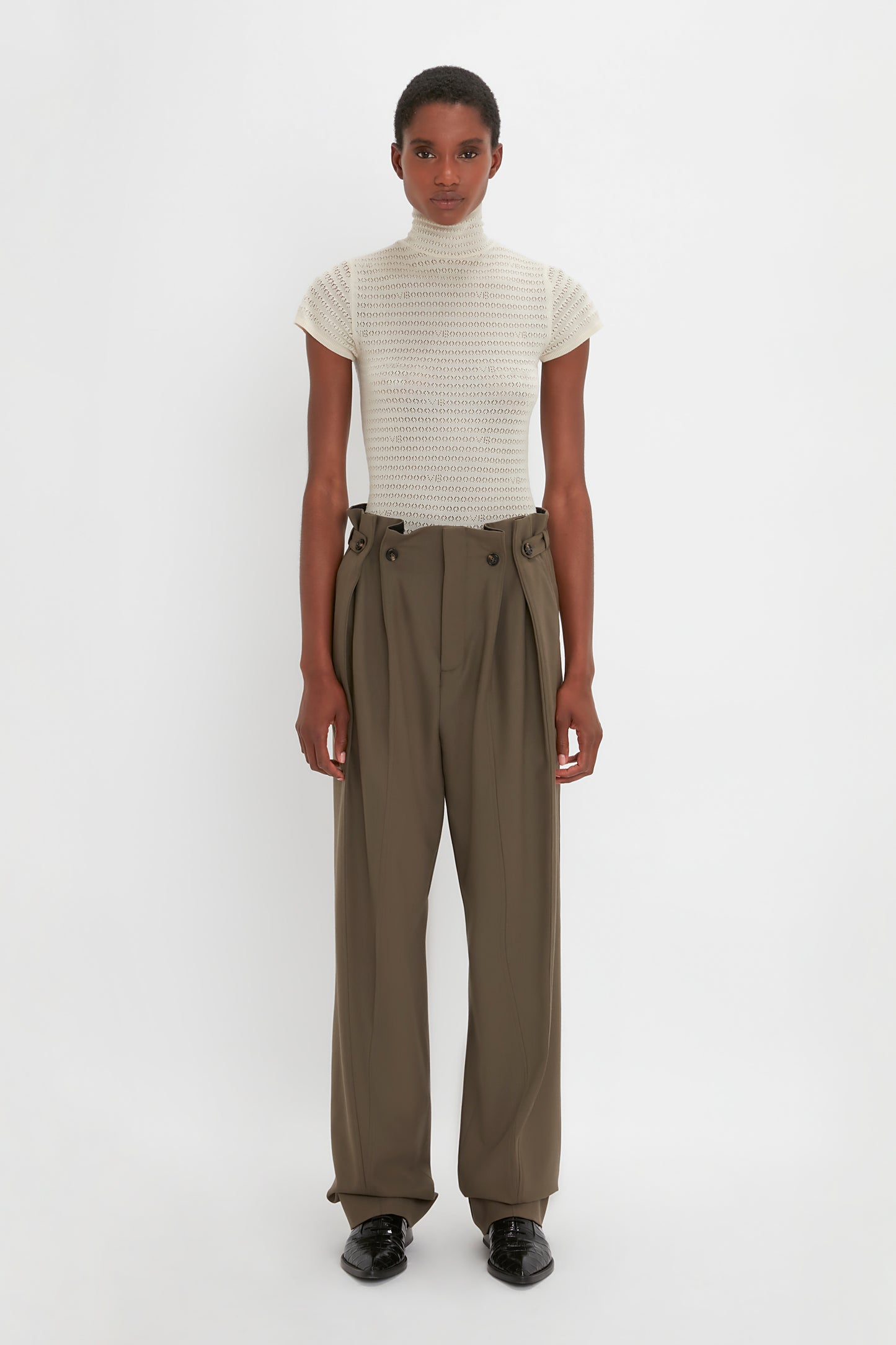 A person stands against a plain white background, wearing a short-sleeve turtleneck top and luxurious Gathered Waist Utility Trouser In Oregano from Victoria Beckham. They also wear dark shoes and have a neutral expression.