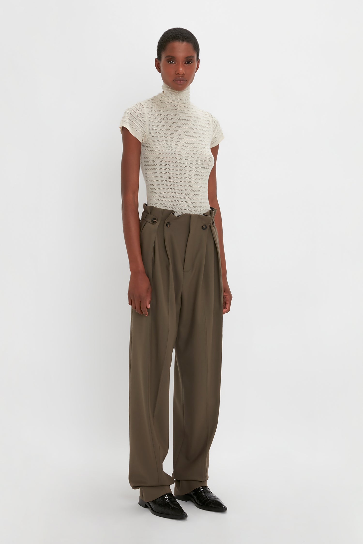 A person stands against a white background, wearing a white short-sleeve turtleneck top and Gathered Waist Utility Trouser In Oregano by Victoria Beckham with an elasticated back waistband. They are also wearing black shoes.