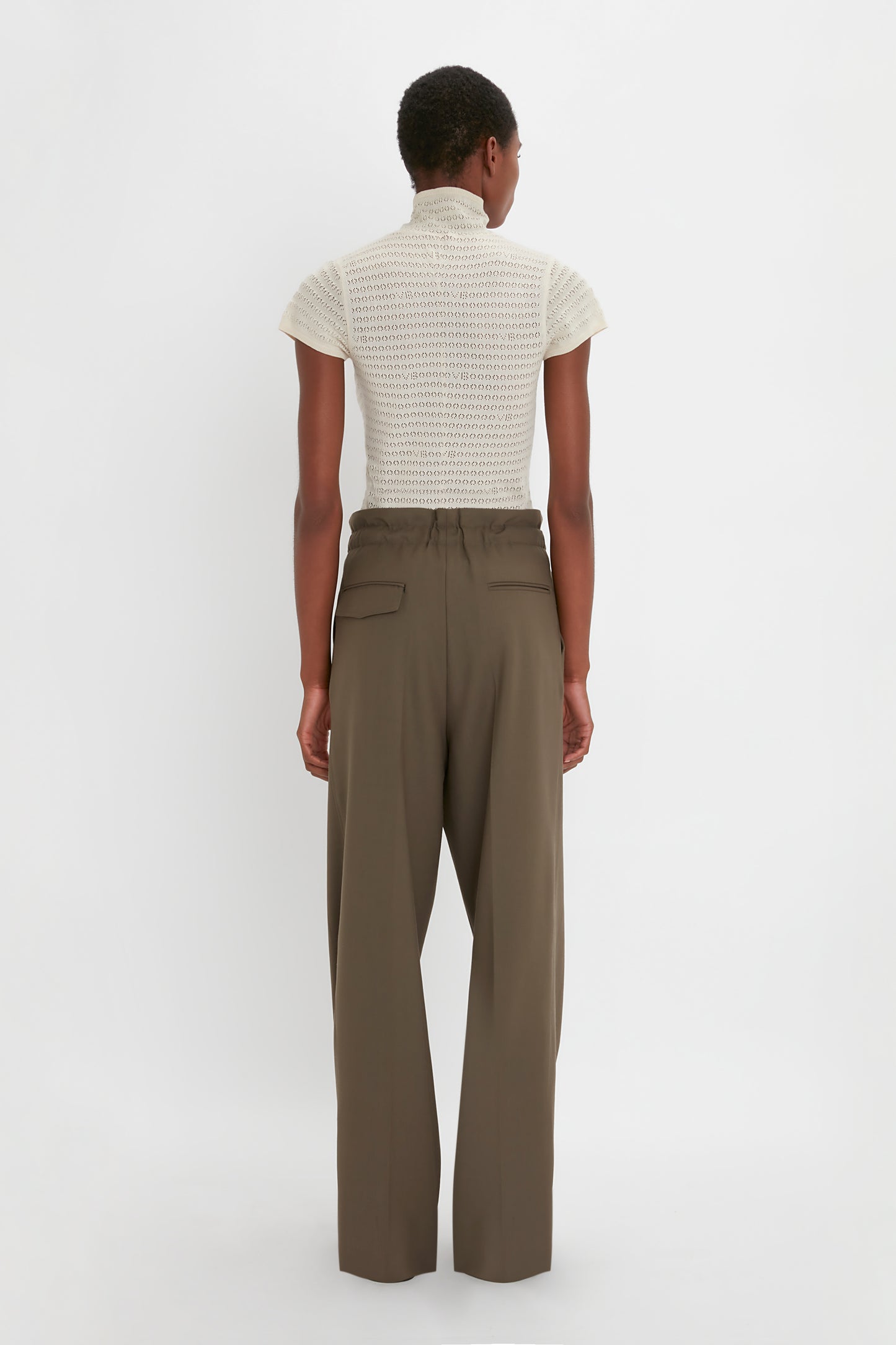 Person standing with their back facing the camera, wearing a short-sleeved knit top and luxurious Gathered Waist Utility Trouser In Oregano by Victoria Beckham. The background is plain white.