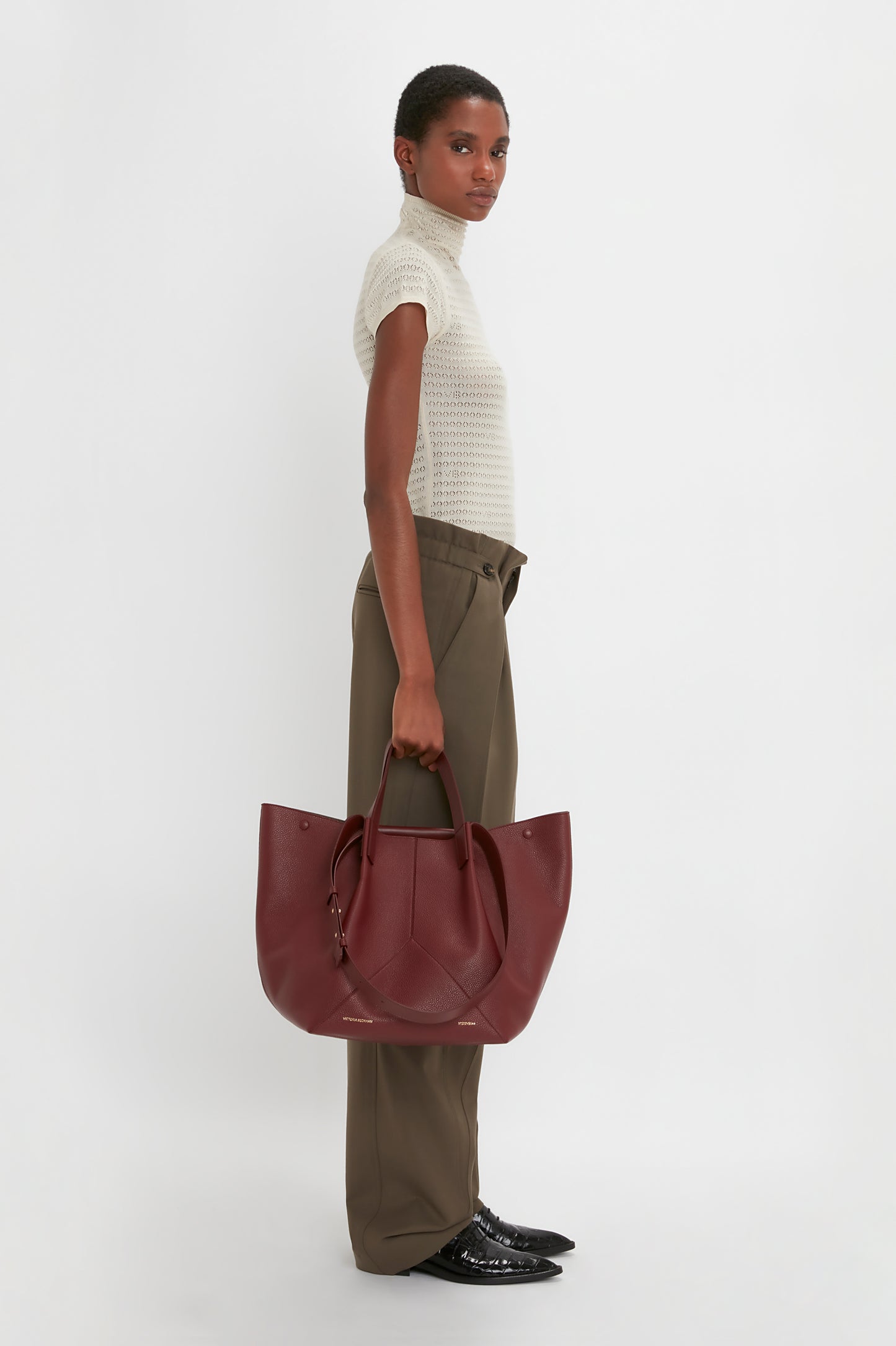 A person stands in profile against a white background, wearing a white turtleneck, luxurious Victoria Beckham Gathered Waist Utility Trouser In Oregano, and black shoes, holding a large maroon handbag.