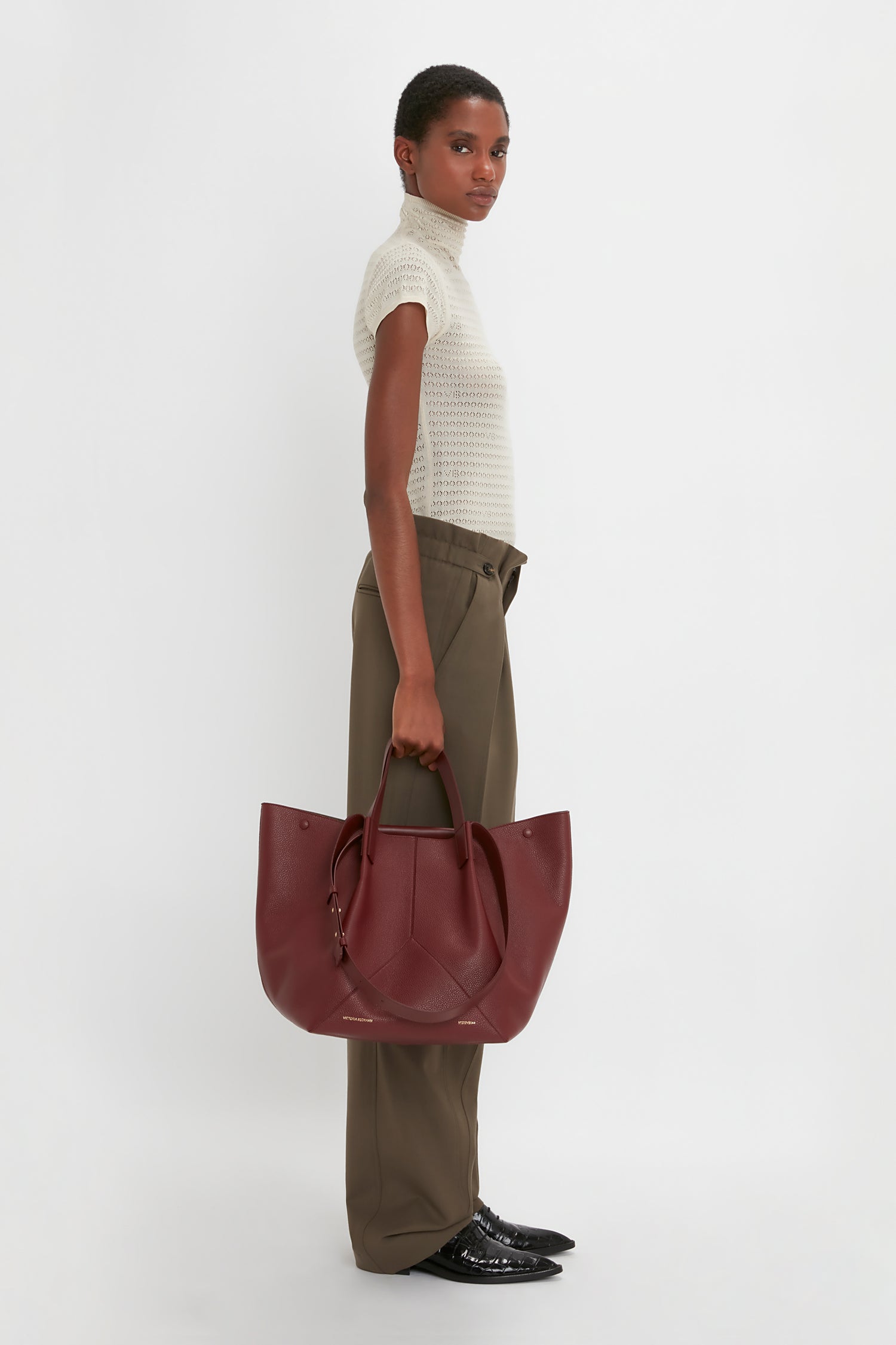 A person stands in profile against a white background, wearing a white turtleneck, luxurious Victoria Beckham Gathered Waist Utility Trouser In Oregano, and black shoes, holding a large maroon handbag.