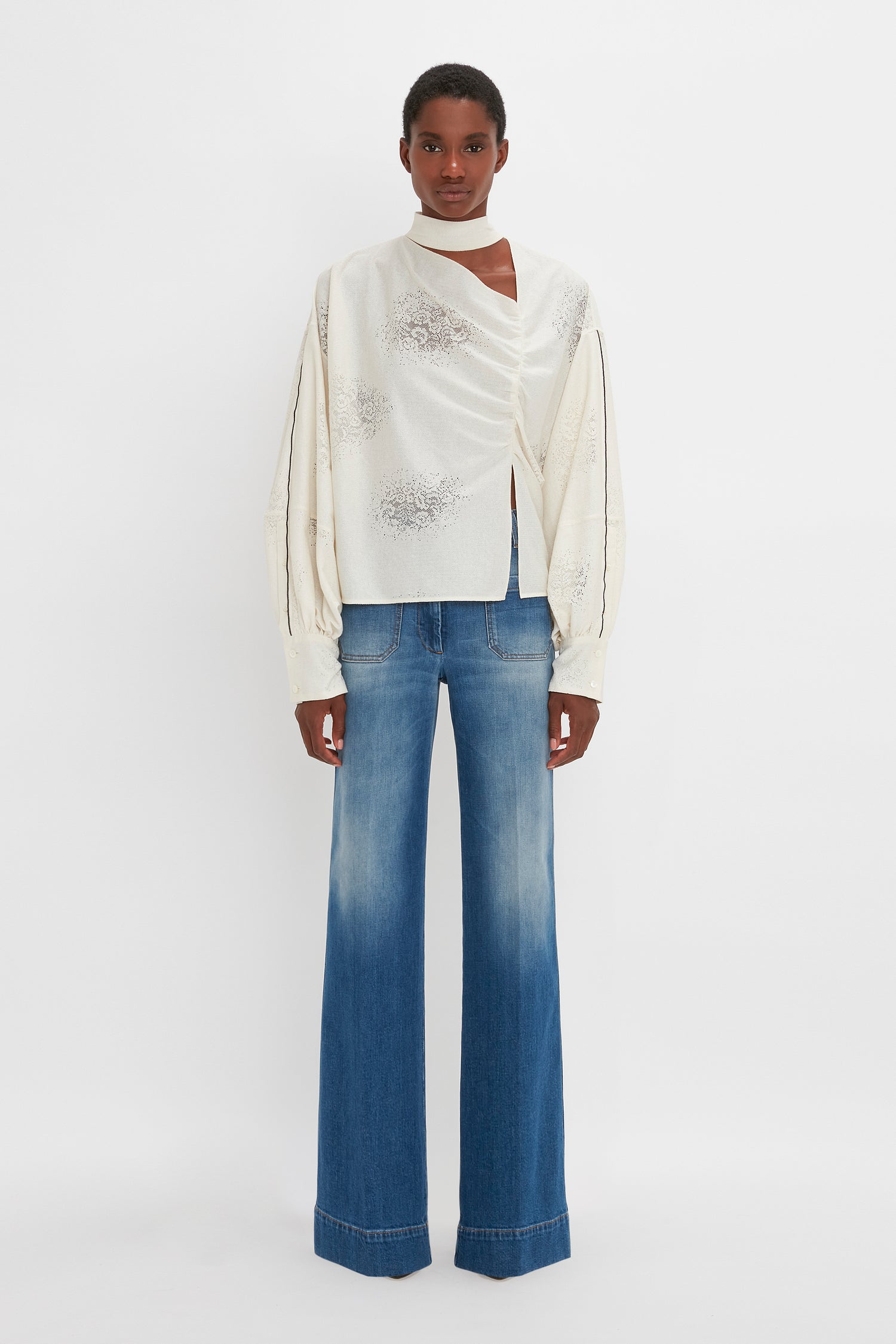 Person wearing a Victoria Beckham Asymmetric Gather Detail Top In Cream paired with faded blue wide-leg jeans, standing against a plain white background.