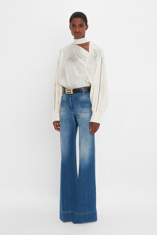 A woman modeling a Victoria Beckham white draped blouse with blouson sleeves and blue flared jeans, posing against a plain white background.