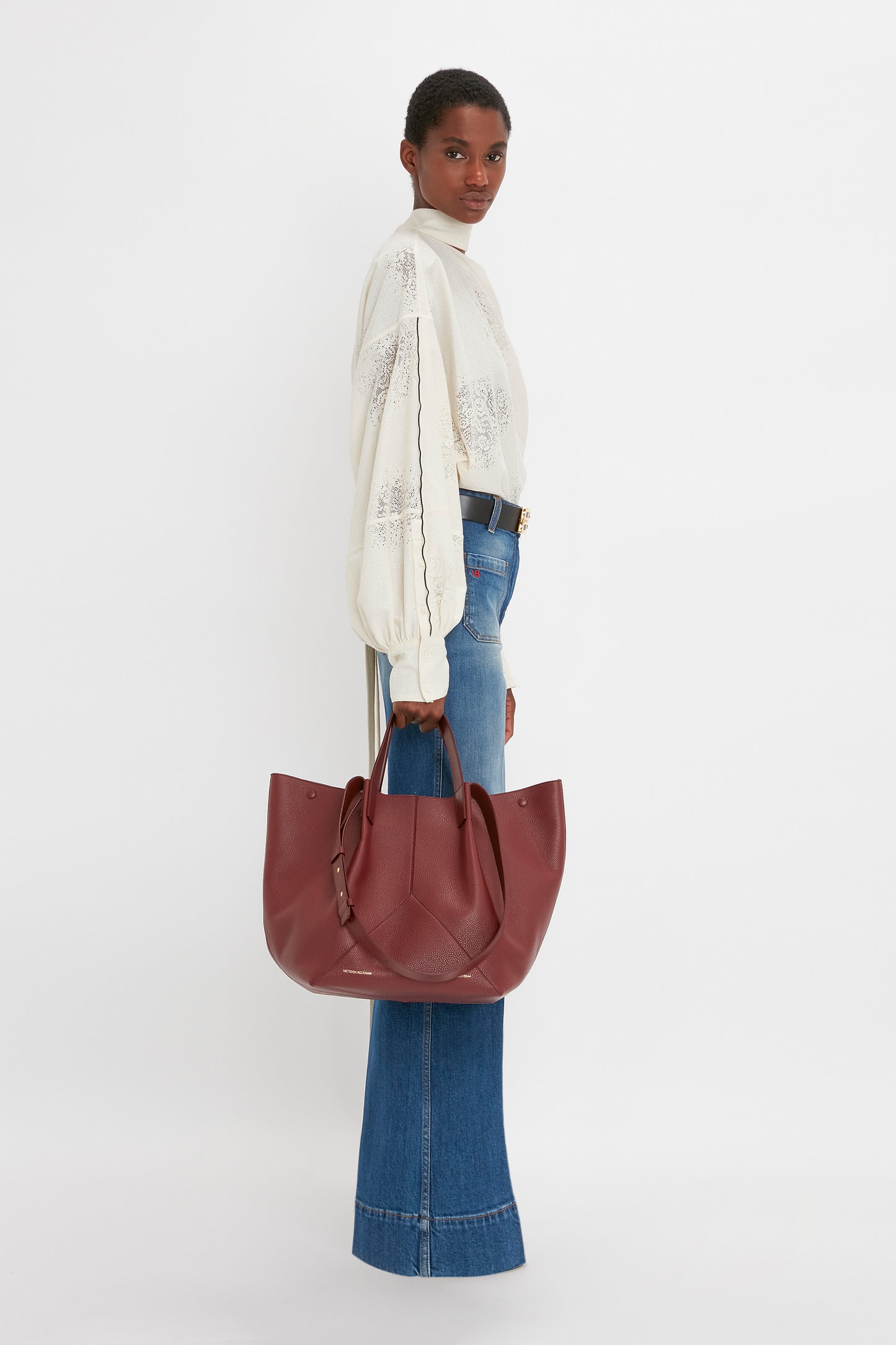 A person stands sideways holding a large maroon handbag, wearing a Victoria Beckham Asymmetric Gather Detail Top In Cream with blouson sleeves and high-waisted wide-leg blue jeans.