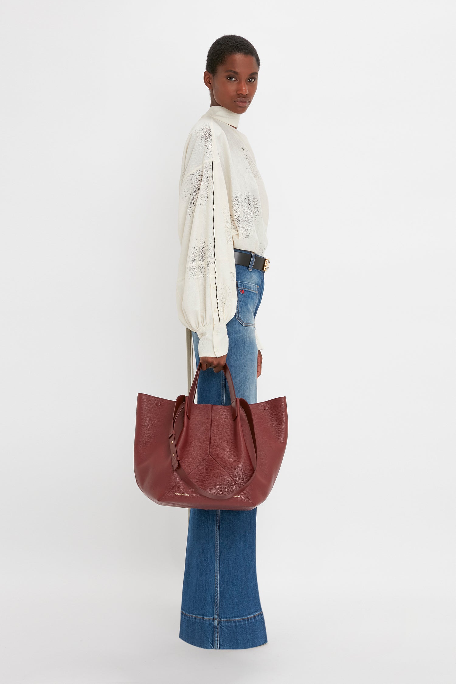 A person stands sideways holding a large maroon handbag, wearing a Victoria Beckham Asymmetric Gather Detail Top In Cream with blouson sleeves and high-waisted wide-leg blue jeans.