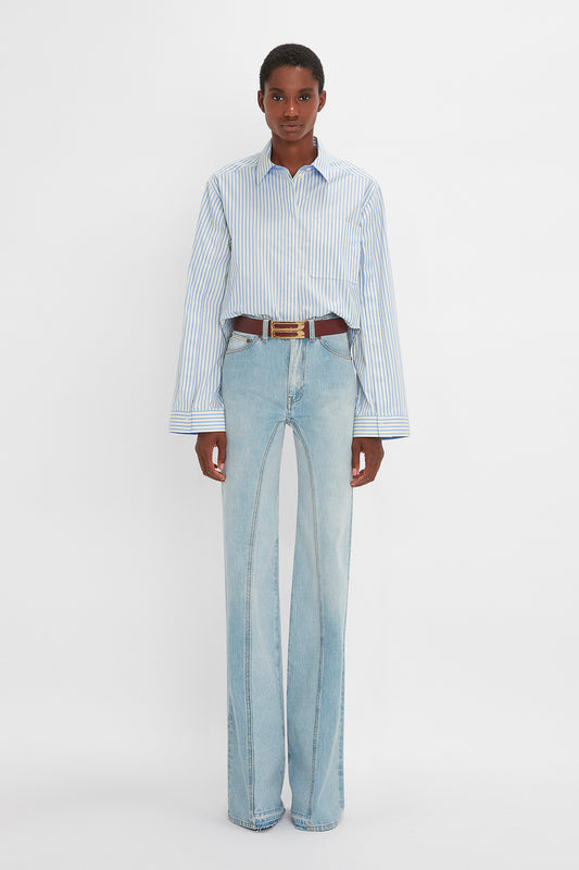 A young man in a vertical-striped blue and white button detail cropped shirt and Victoria Beckham Bianca Jean In Light Blue Denim stands facing the camera on a plain background.