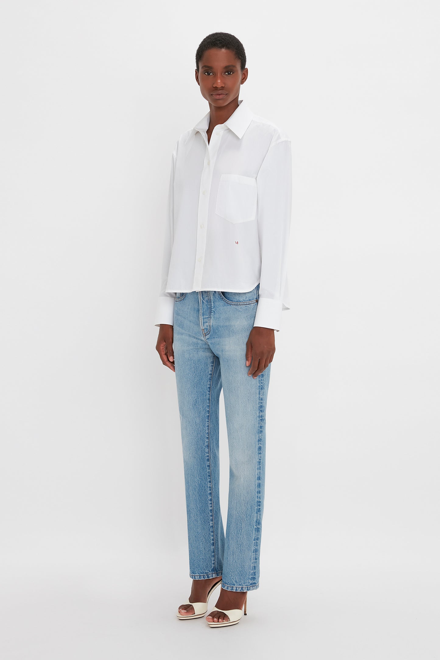 A black woman in a Victoria Beckham classic white shirt and blue jeans, standing against a plain white background. She is wearing silver heels.