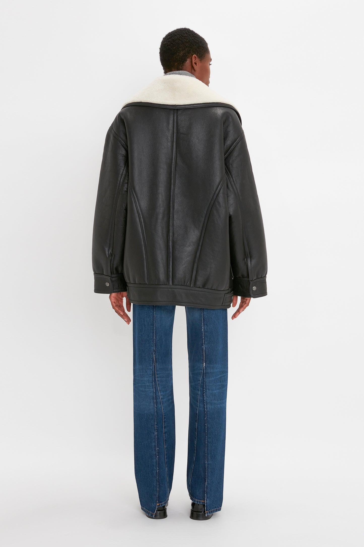 A person wearing a black leather jacket with a white fur collar, blue jeans, and black shoes stands and faces away from the camera against a plain white background, evoking the timeless style of the Shearling Coat In Monochrome by Victoria Beckham.