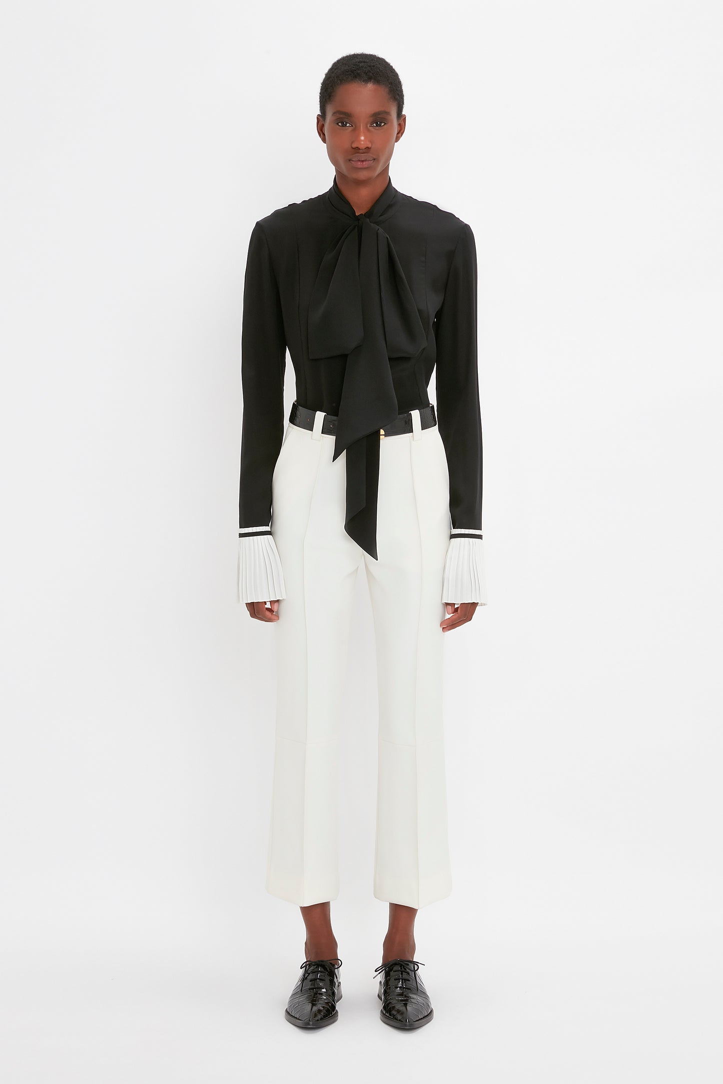 A person stands against a plain white background, wearing a black blouse with a large bow and pleated cuffs, white tailored pants, and Victoria Beckham's Pointy Toe Flat Lace Up In Black Croc-Effect Leather.