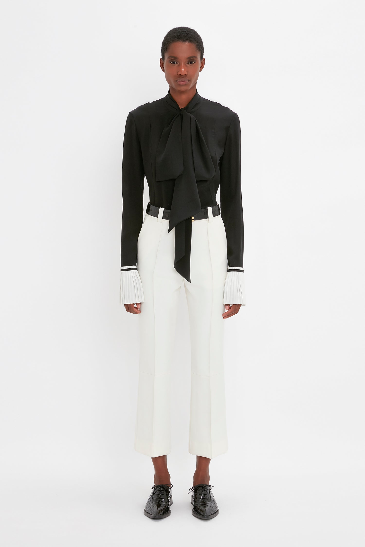 A person stands against a plain white background, wearing a black blouse with a large bow and pleated cuffs, white tailored pants, and Victoria Beckham's Pointy Toe Flat Lace Up In Black Croc-Effect Leather.